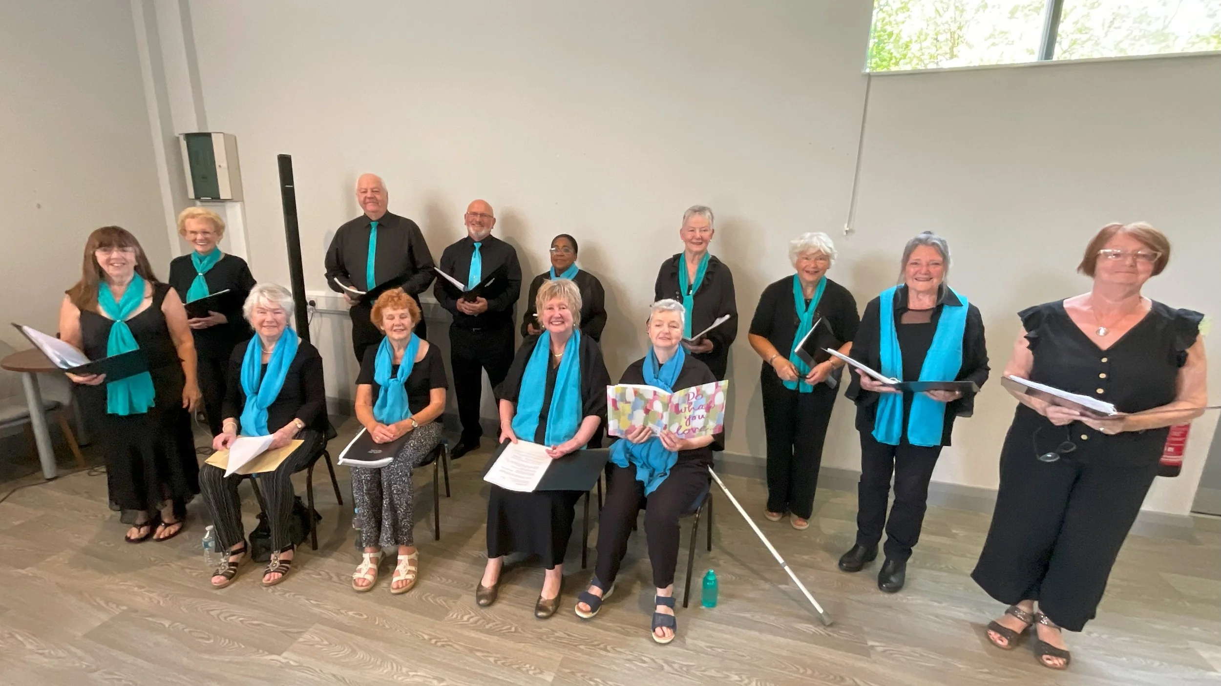 A group of elderly choir singers posing together indoors, wearing black clothing with teal scarves, holding music sheets, with some standing and some seated.