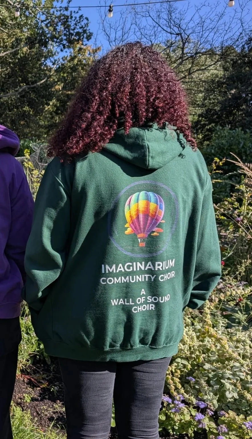 Person with curly hair wearing a green hoodie with a colorful hot air balloon graphic and the words 'Imaginarium Community Choir, A Wall of Sound Choir' on the back, standing outdoors among bushes and trees.