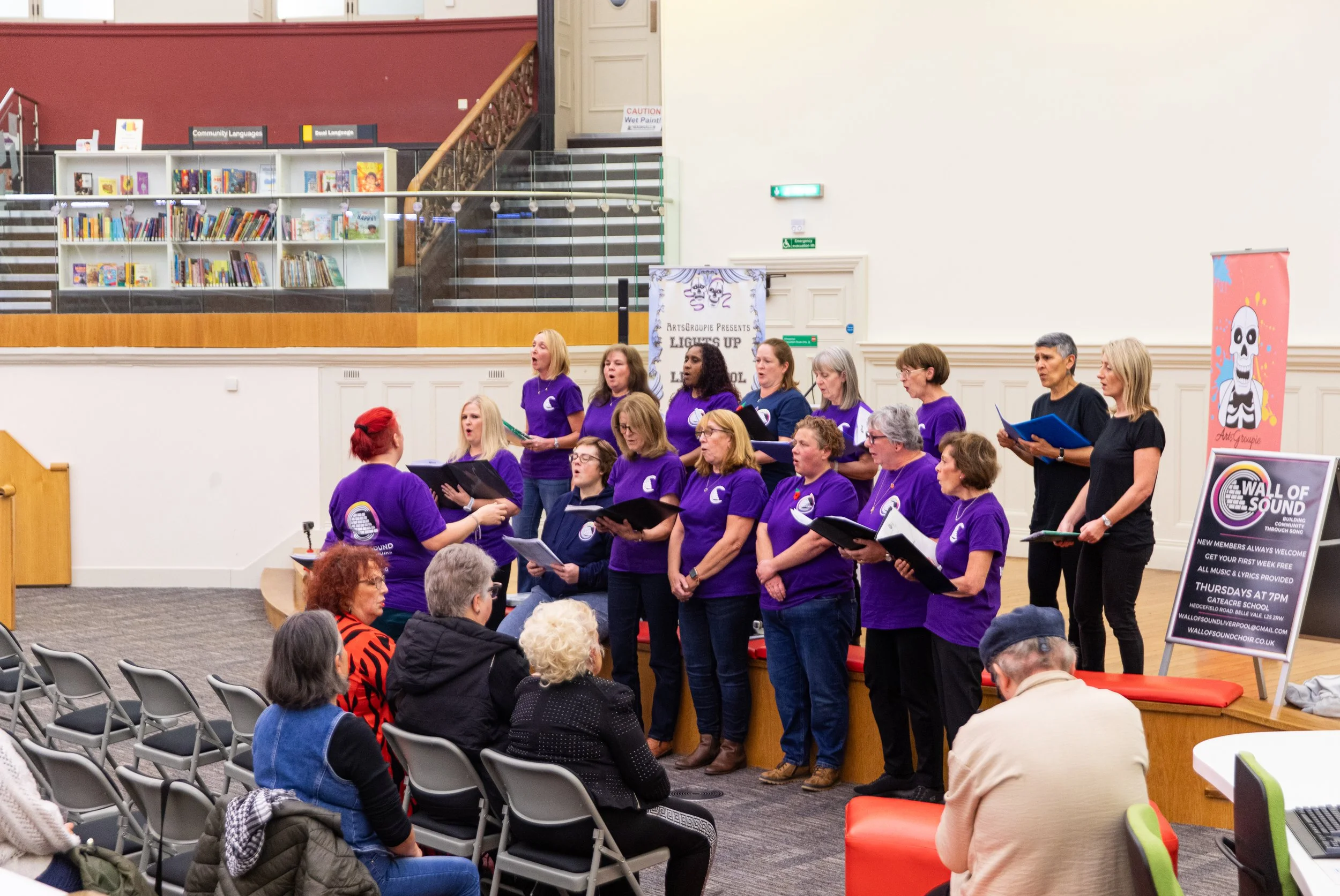 A choir of adults performing on a stage in a library, wearing matching purple shirts with a crescent moon logo, while an audience of diverse individuals watches.