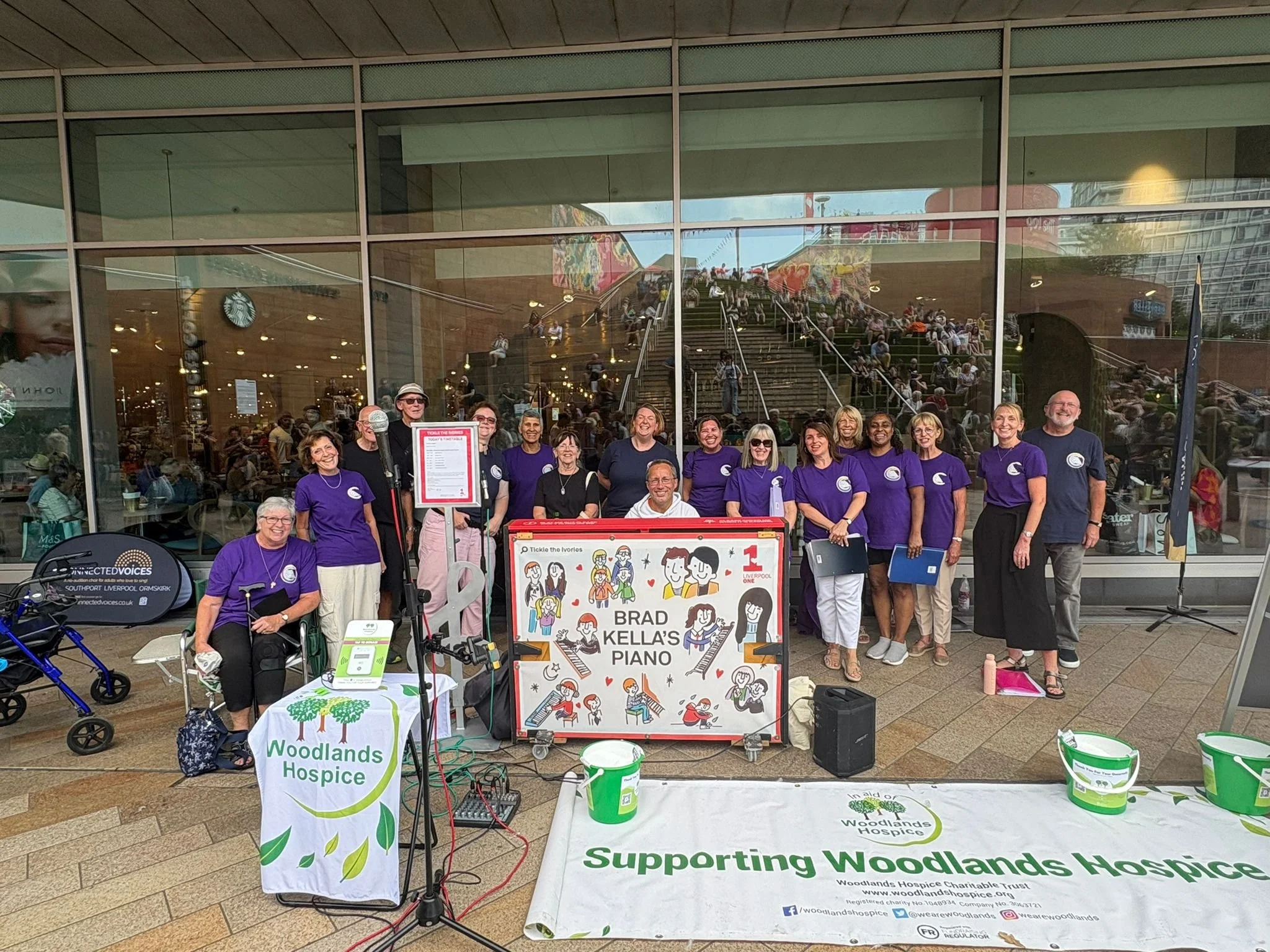 Group of people standing outside a building supporting Woodlands Hospice. Some hold clipboards, and there is a decorated sign that reads 'Brad Kella's Piano'. A booth with a Woodlands Hospice banner is in front, and a crowd is visible through the glass behind.
