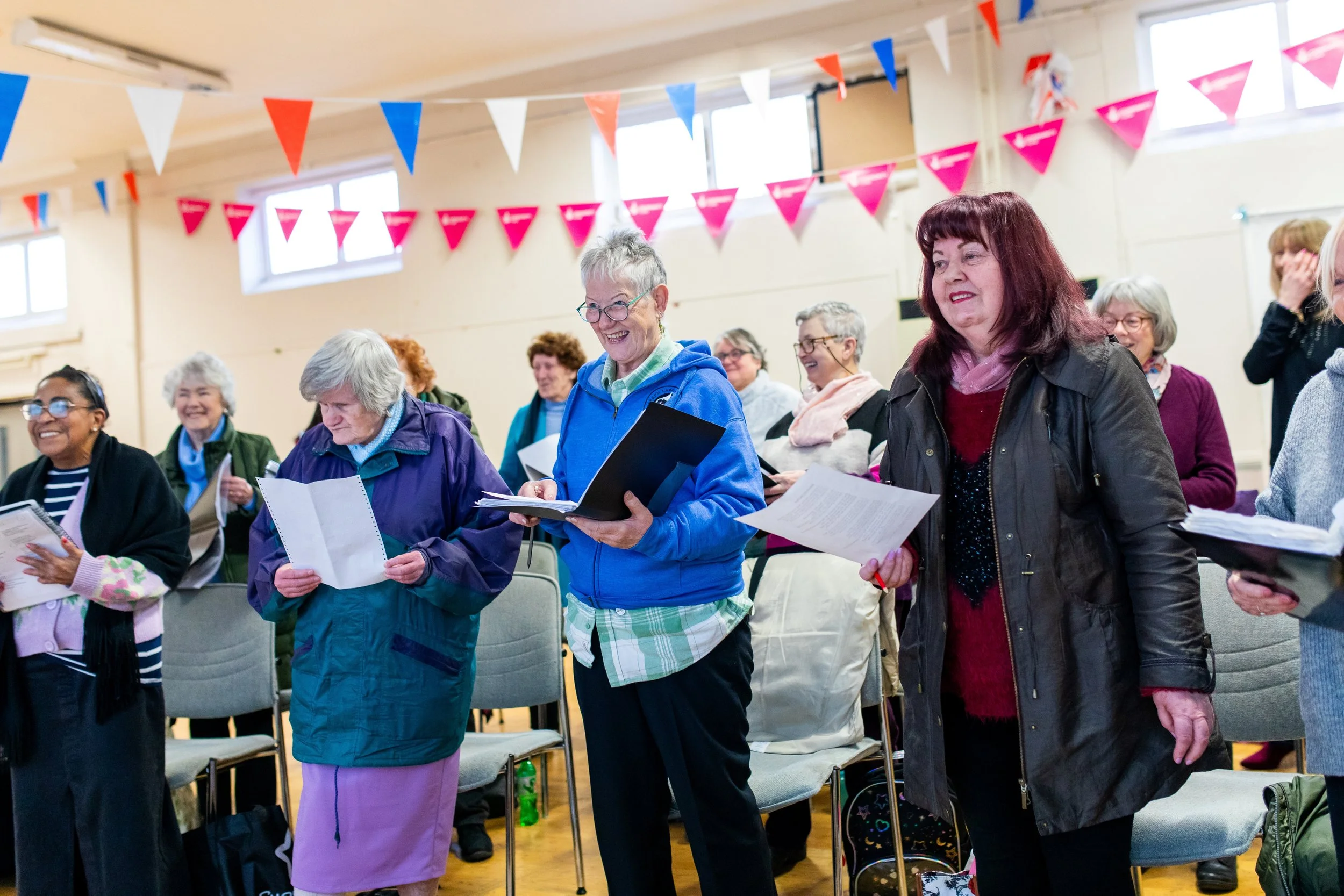 Group of women in a community hall, standing and singing, holding songbooks, with red, white, and blue triangular flags hanging from the ceiling.