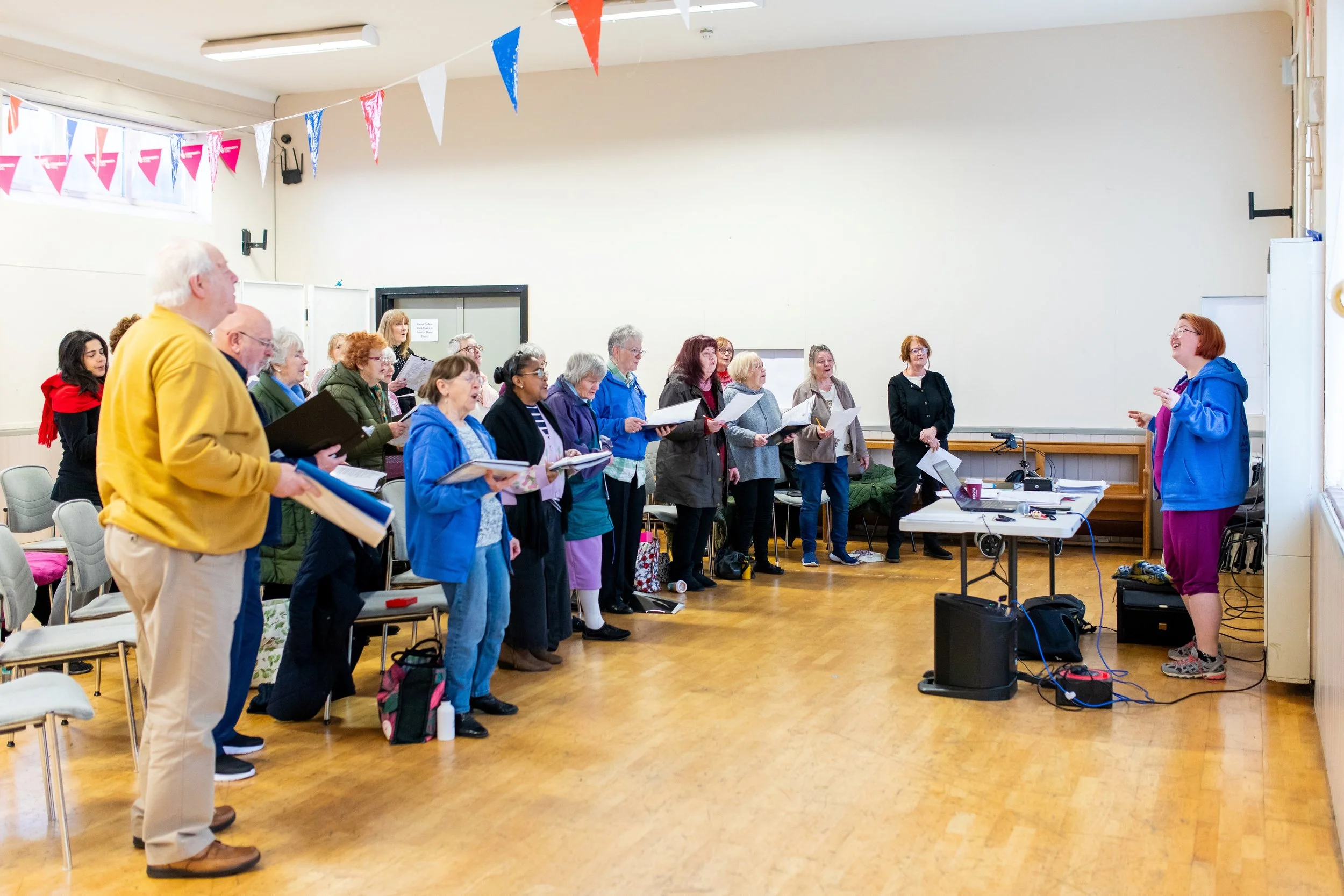 A choir practicing in a room with an instructor standing in front of them. The group is composed of elderly and middle-aged individuals holding sheet music. The instructor is wearing a blue hoodie and purple pants, standing next to a table with a laptop and sound equipment. There are festive decorations with pink, blue, and white bunting hanging from the ceiling.