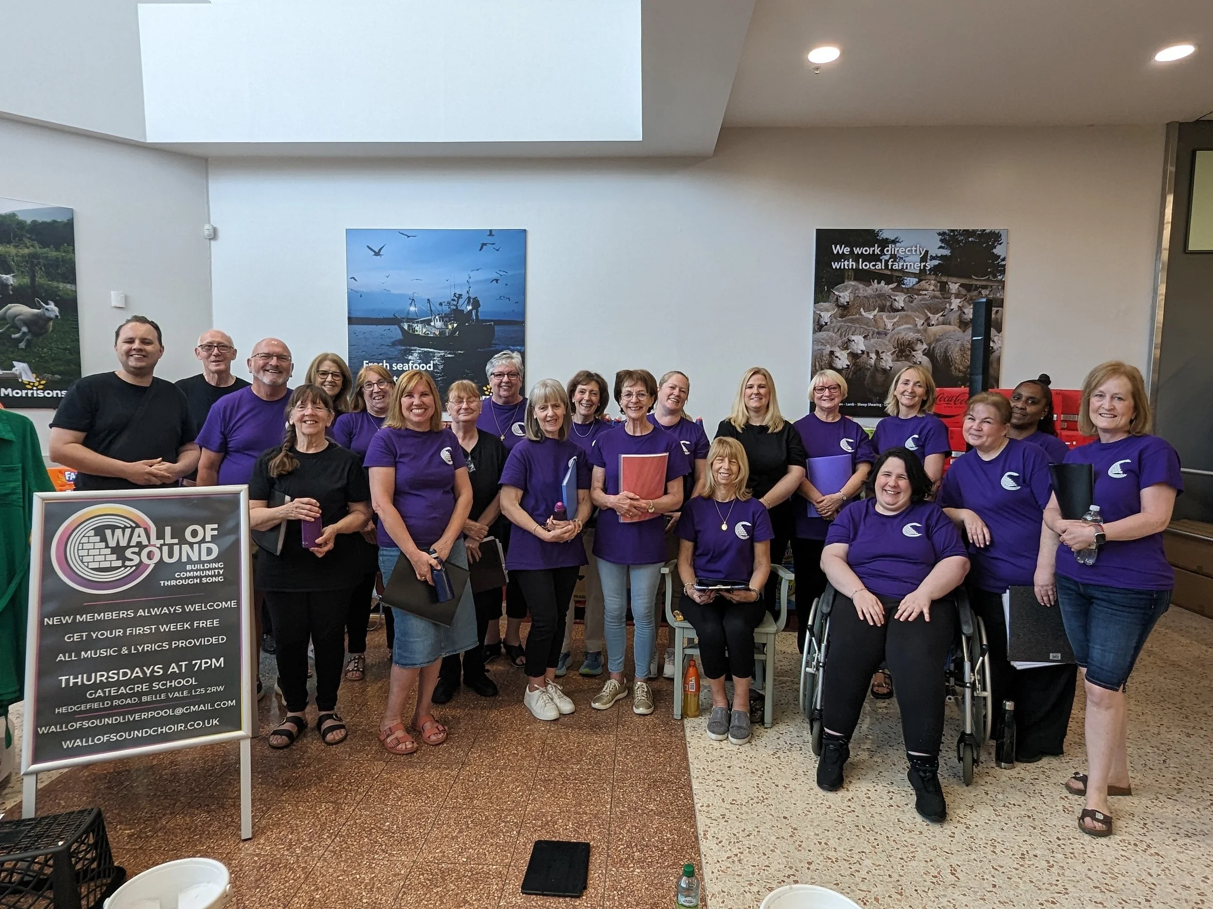 Group of people from the Wall of Sound choir smiling and standing together indoors, some holding folders or water bottles, with posters and signs about the choir and their activities in the background.