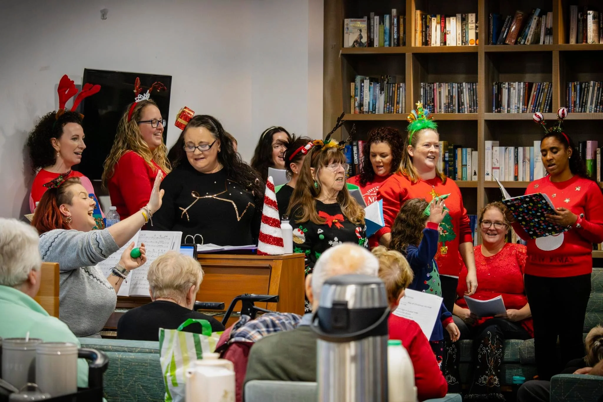 A group of people singing Christmas carols during a holiday gathering, some wearing festive sweaters and reindeer antlers, in a room with a bookshelf in the background and an audience seated at tables.