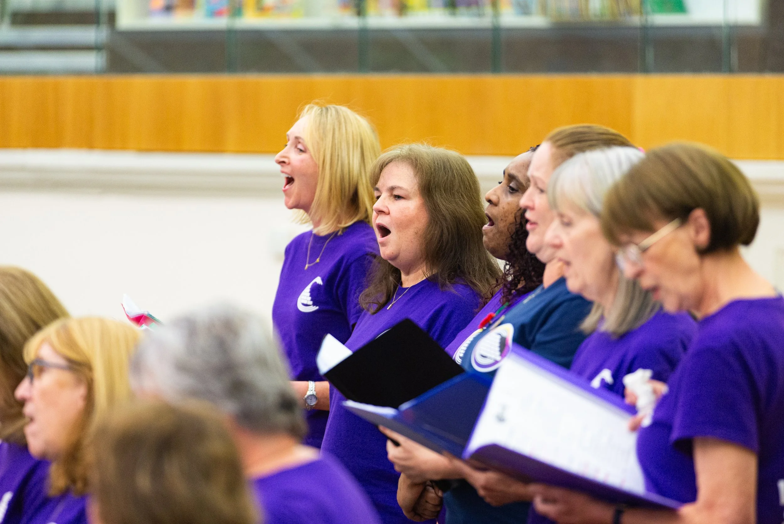 Group of women singing in a choir, wearing purple shirts, holding folders with sheet music, in a large room with wood-paneled walls.