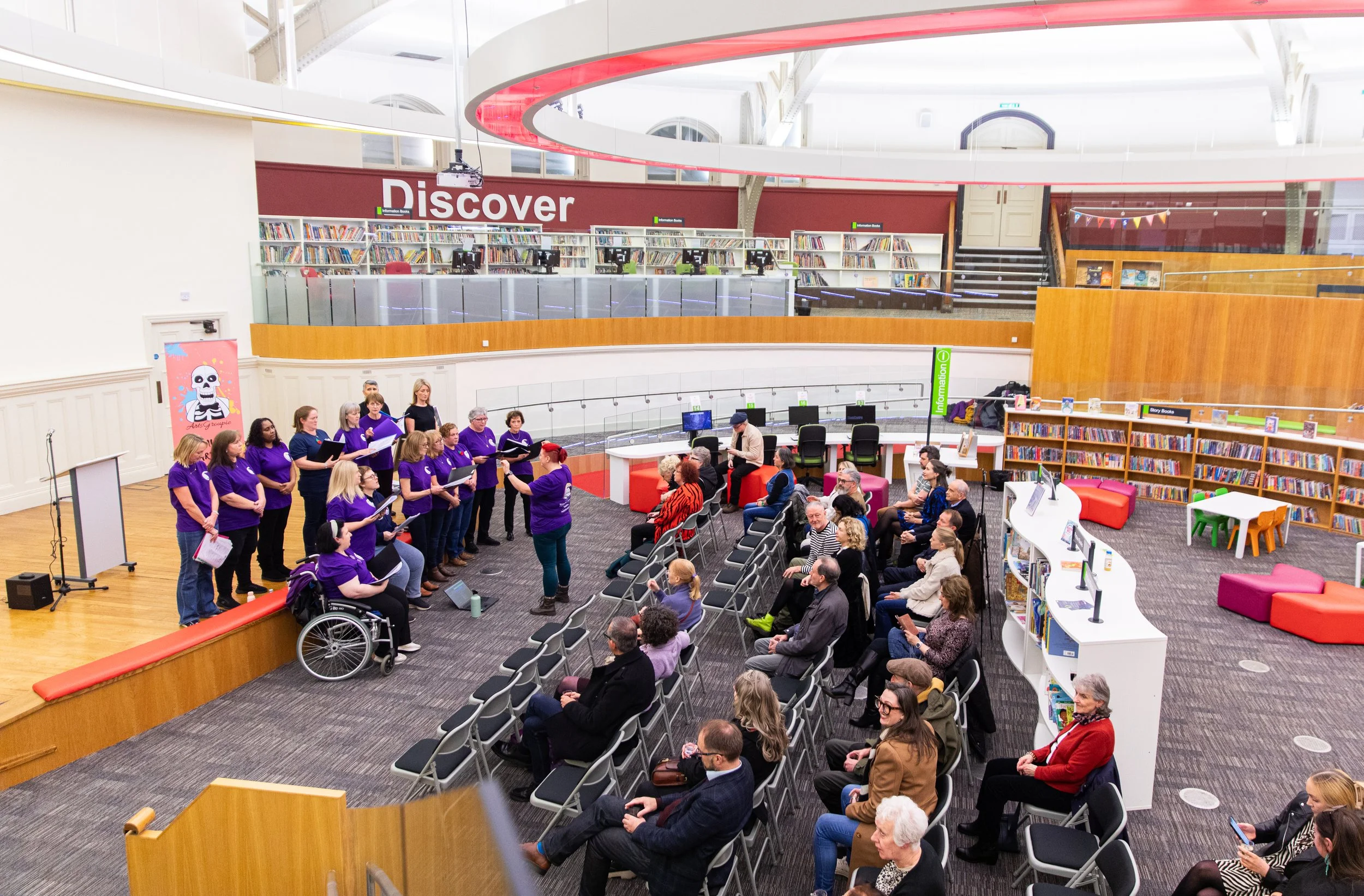 A choir performance in a modern library's event space; choir members wearing purple shirts, some in wheelchairs, singing to an audience seated in chairs. Audience mingles and watches attentively. Shelves of books and a children's area are visible in the background.