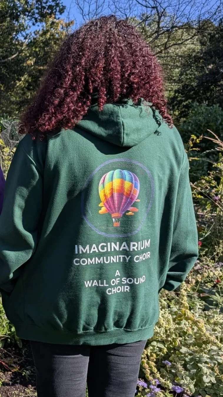 A person with curly hair wearing a green hoodie with a rainbow-colored hot air balloon graphic and text promoting the Imaginarium Community Choir, standing outdoors among plants and trees.