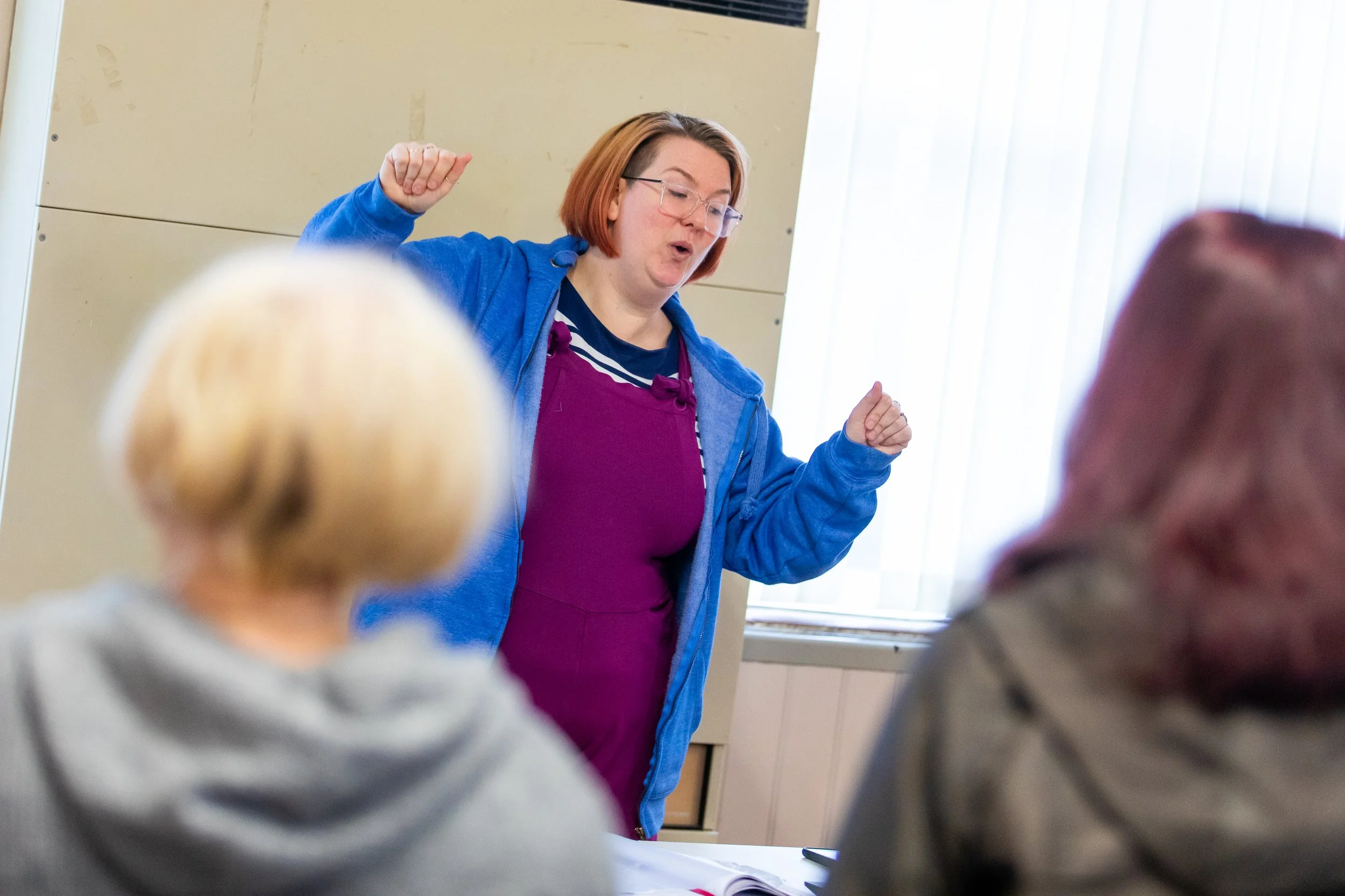A woman with glasses and shoulder-length hair, wearing a purple dress and blue jacket, appears to be teaching or speaking passionately to a group of people in a classroom. The group includes at least two people visible from behind, one with blonde hair and the other with reddish-brown hair.
