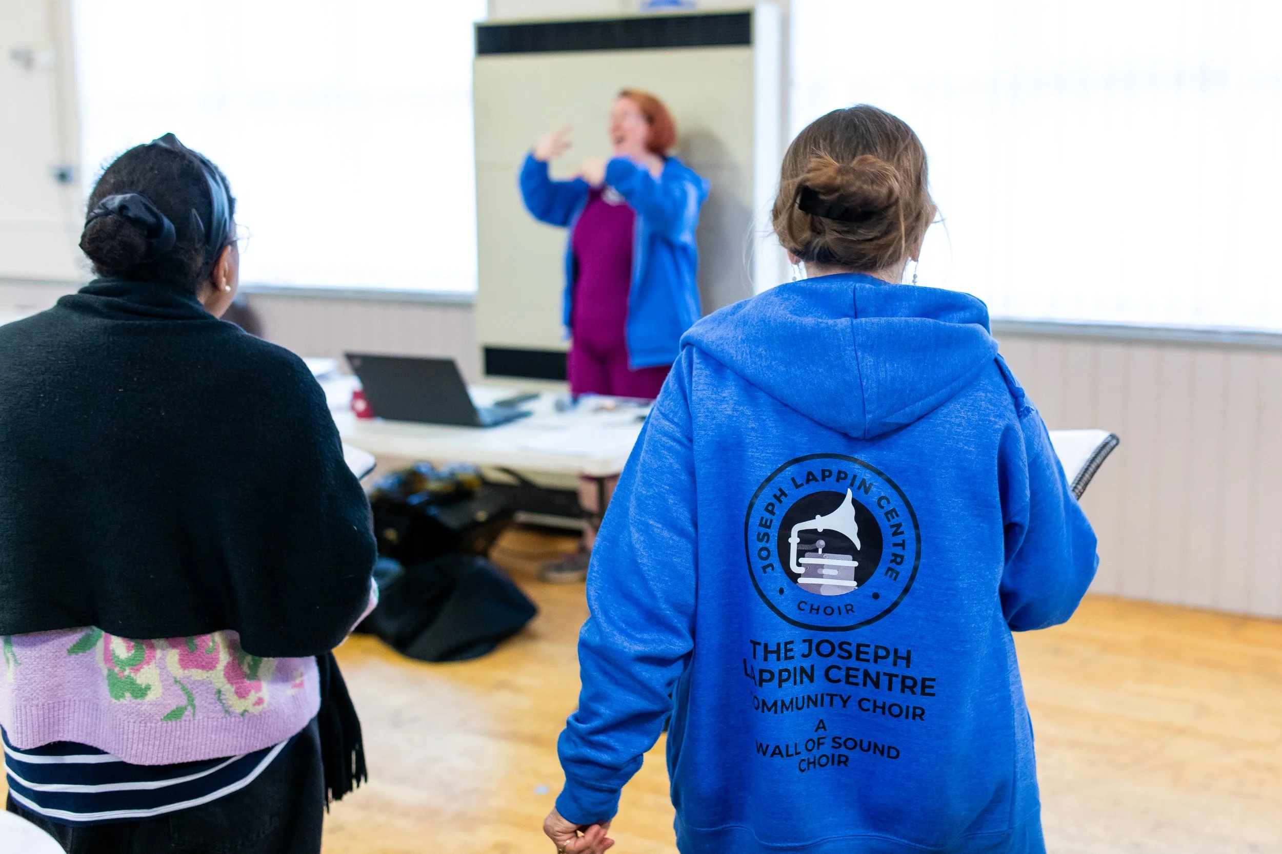 A choir practice takes place with a woman conducting and two women facing her. One woman wears a black sweater with a pink scarf, and the other wears a blue hoodie with a choir logo and text on the back.