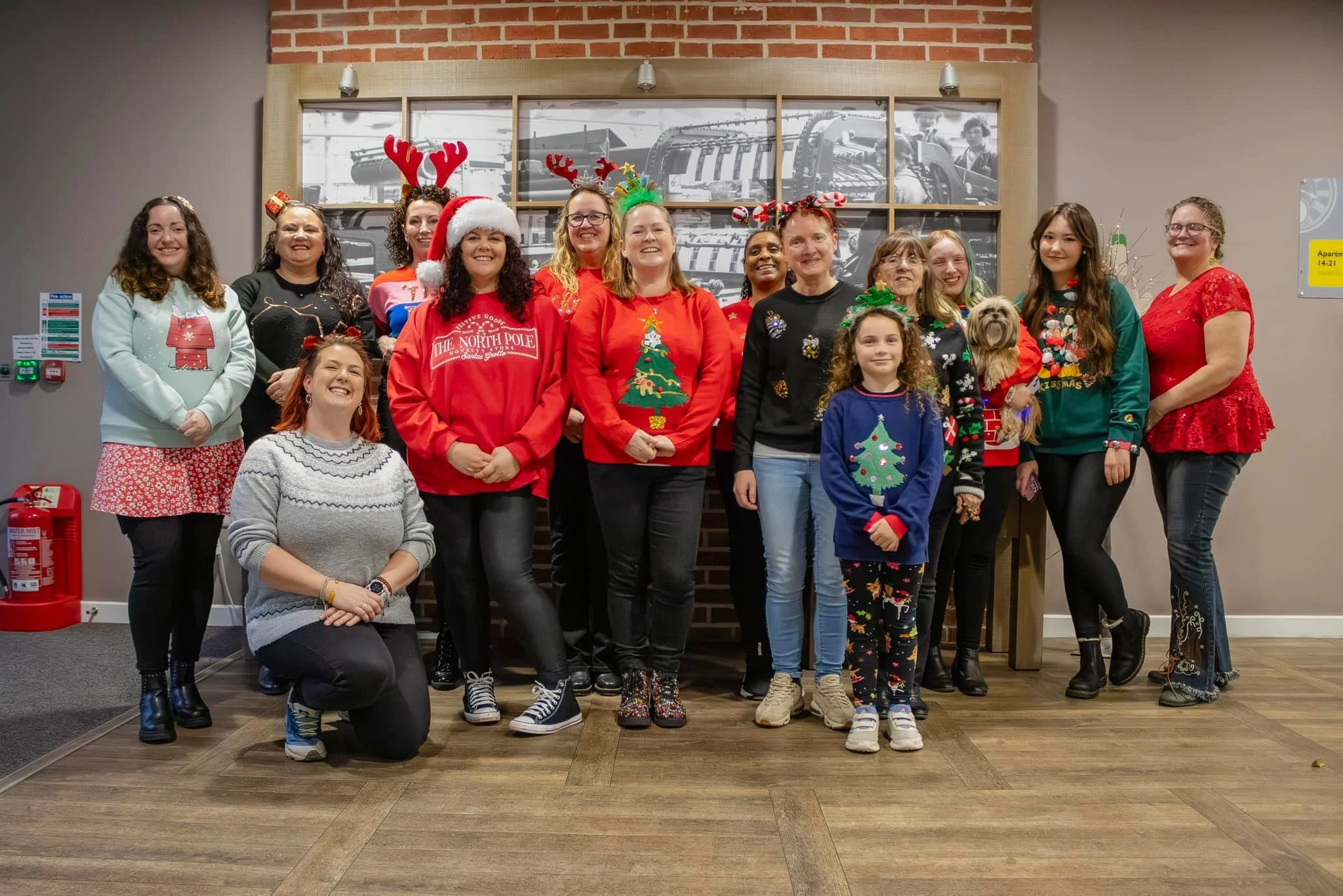 Group of women and girls celebrating Christmas, wearing festive sweaters and accessories like reindeer antlers and Christmas hats, in an indoor setting with a brick wall and black-and-white framed photos in the background.