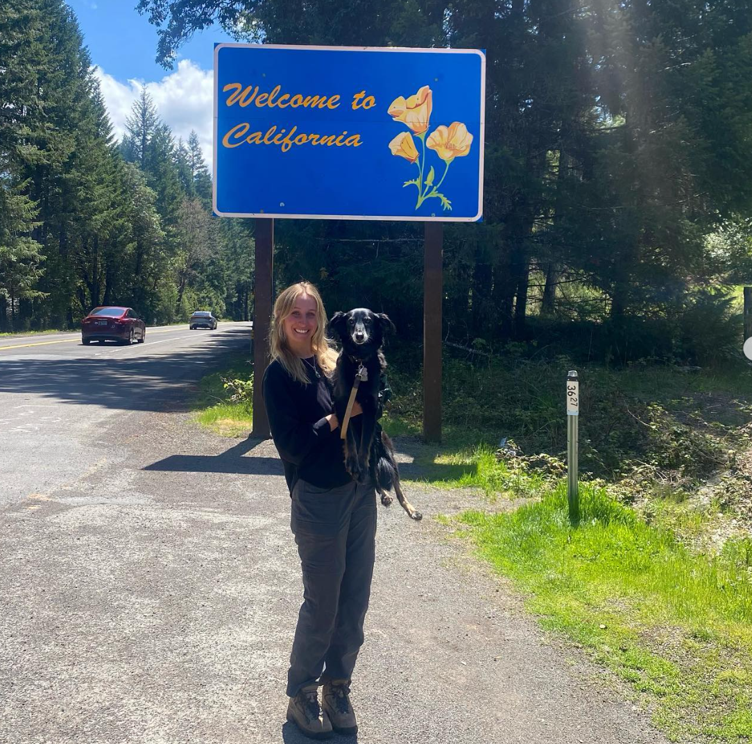 A woman holding a black dog standing on the side of a road in front of a "Welcome to California" sign surrounded by trees and blue sky.