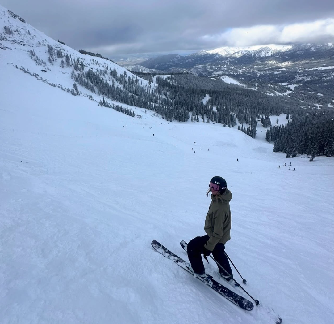 A person skiing on a snowy mountain slope with a scenic view of snow-covered mountains and pine trees in the background.