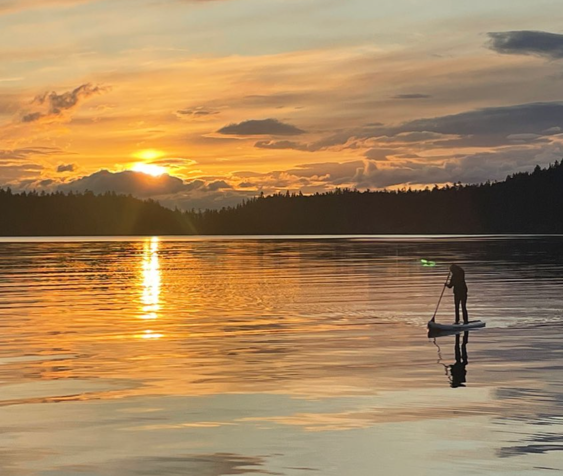 A person paddleboarding on a calm lake during sunset with a colorful sky, distant treeline, and reflections on the water.