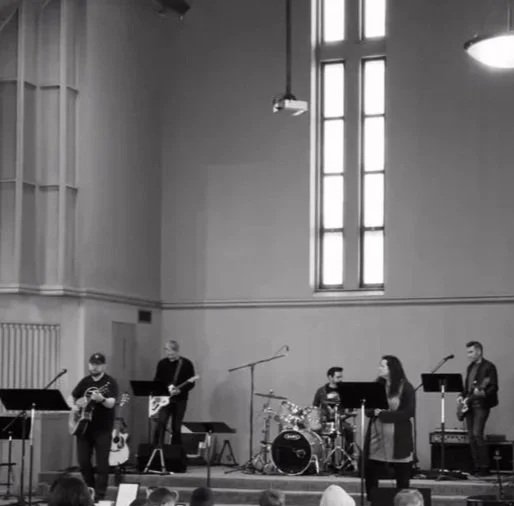 A band in Minneapolis performing on stage inside a church, with five members playing guitars, drums, and singing. Audience watching the performance.