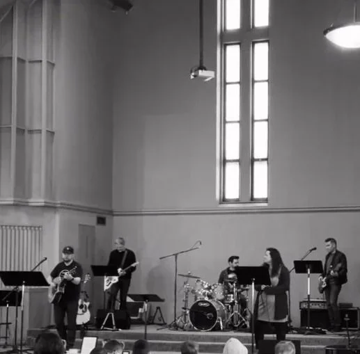 A band performing on stage inside a church in South Minneapolis with a large window and a projection screen, with an audience seated in front.