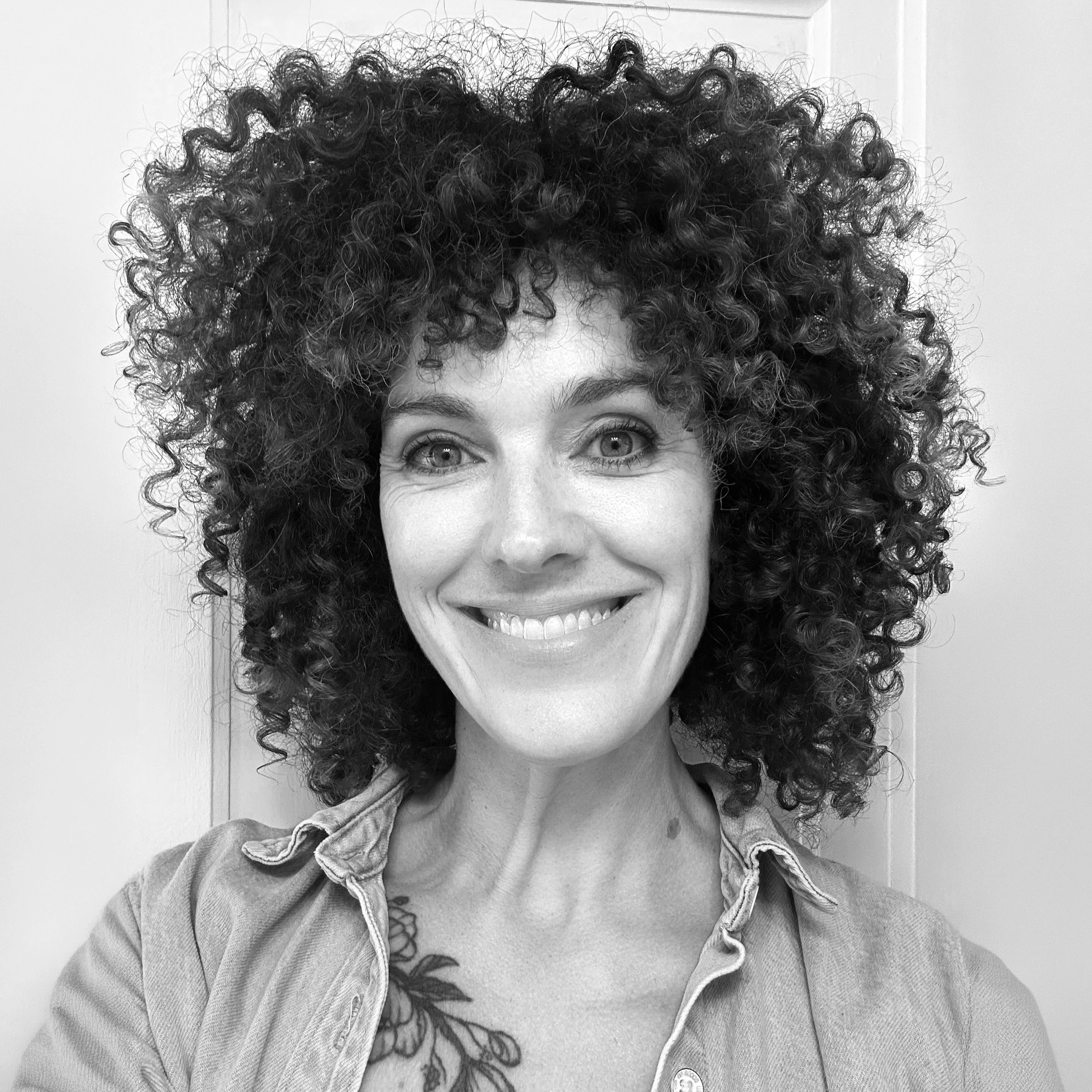 Black and white portrait of a smiling woman with short curly hair, wearing earrings and layered necklaces, against a plain background.