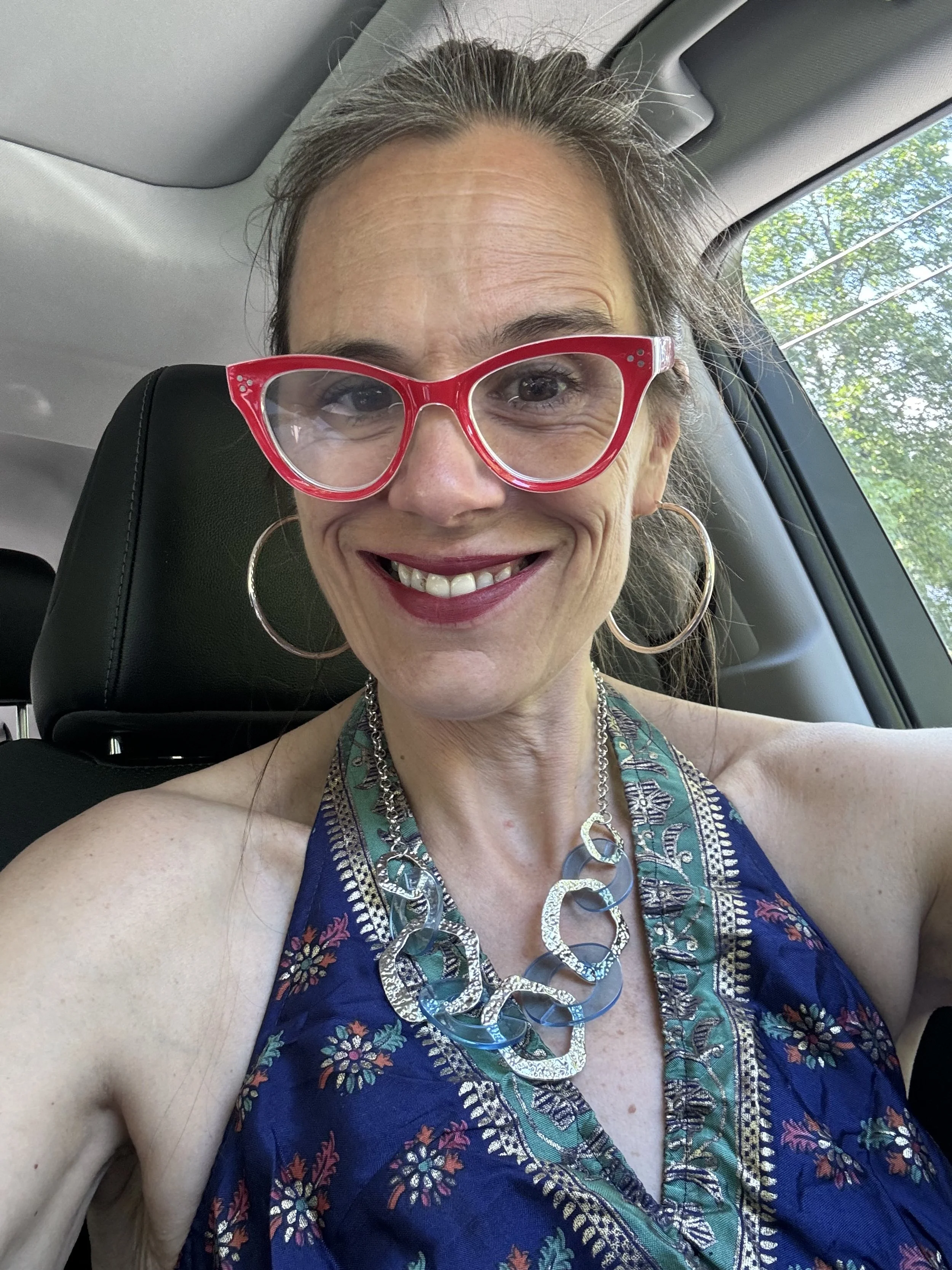 A woman smiling in a car, wearing large pink glasses, hoop earrings, a colorful patterned dress, and a chunky silver necklace.
