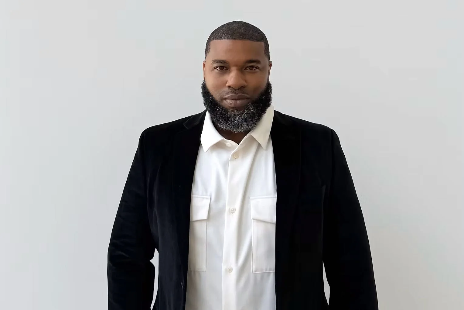 A man with a beard wearing a white shirt and black blazer standing against a plain light gray wall.