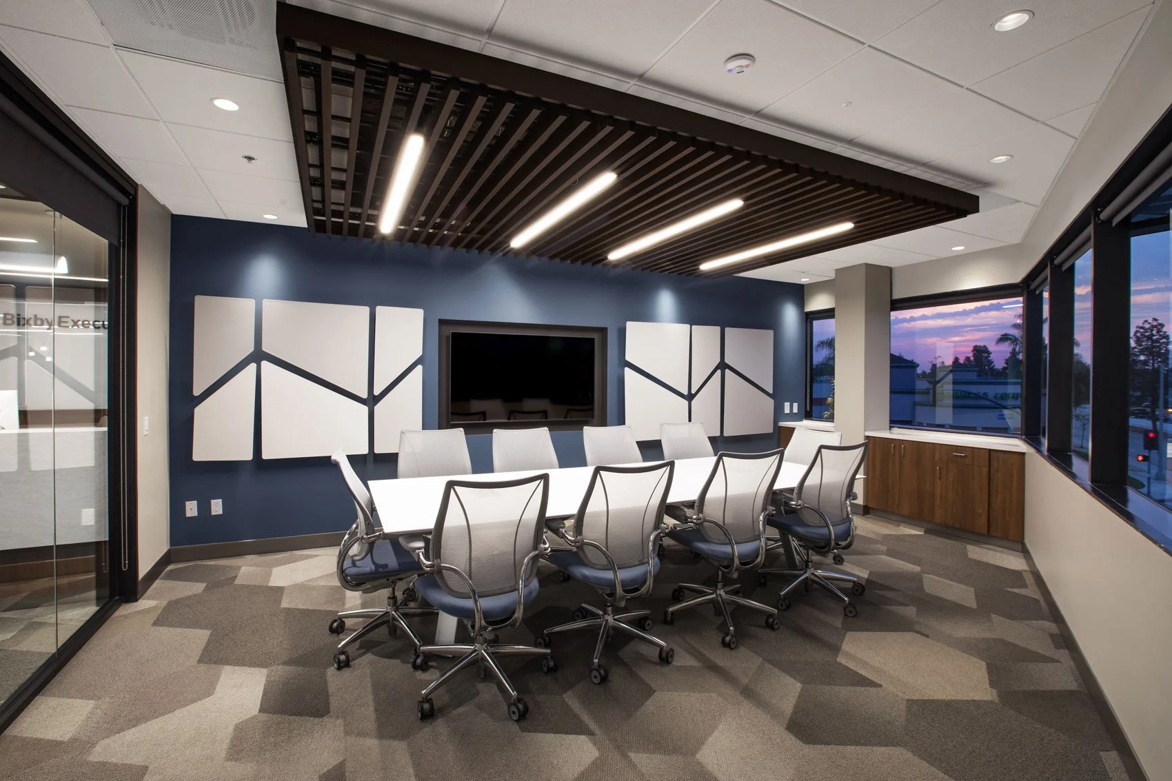 Modern conference room with a long white table, nine white chairs, a blue accent wall with sound panels, a large flat screen TV, and big windows showing a sunset outside.