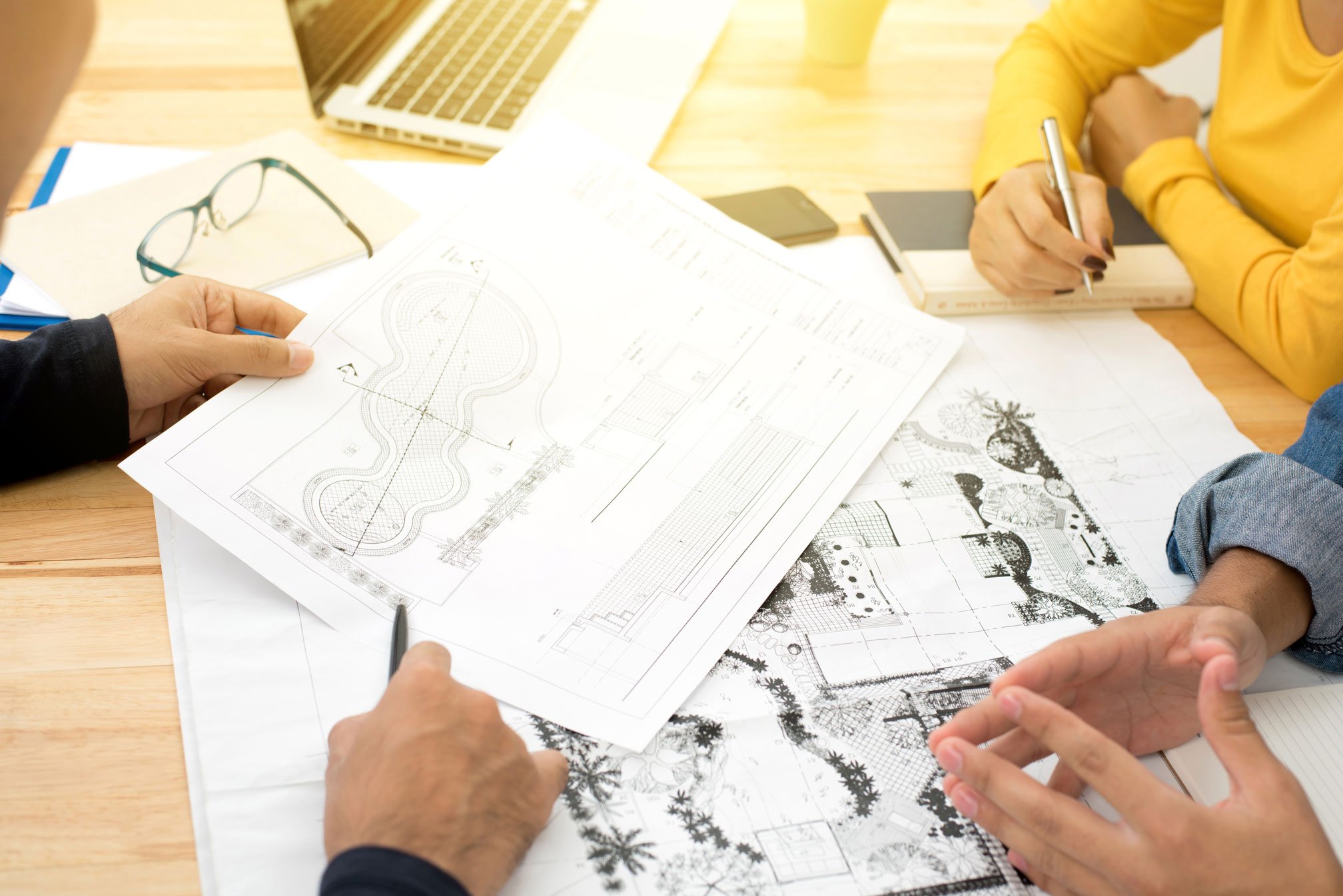 People reviewing landscape architecture plans on a wooden table. Blueprints, notes, a laptop, eyeglasses, and notebooks are visible.