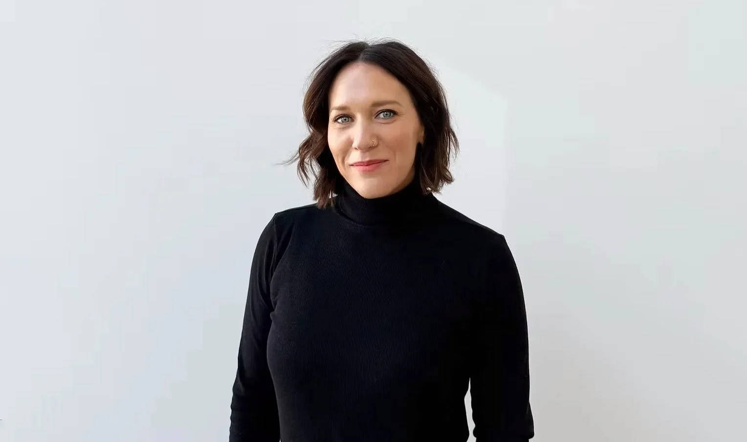 A woman with shoulder-length brown hair, wearing a black turtleneck sweater, standing against a plain light gray background, smiling slightly, facing the camera.