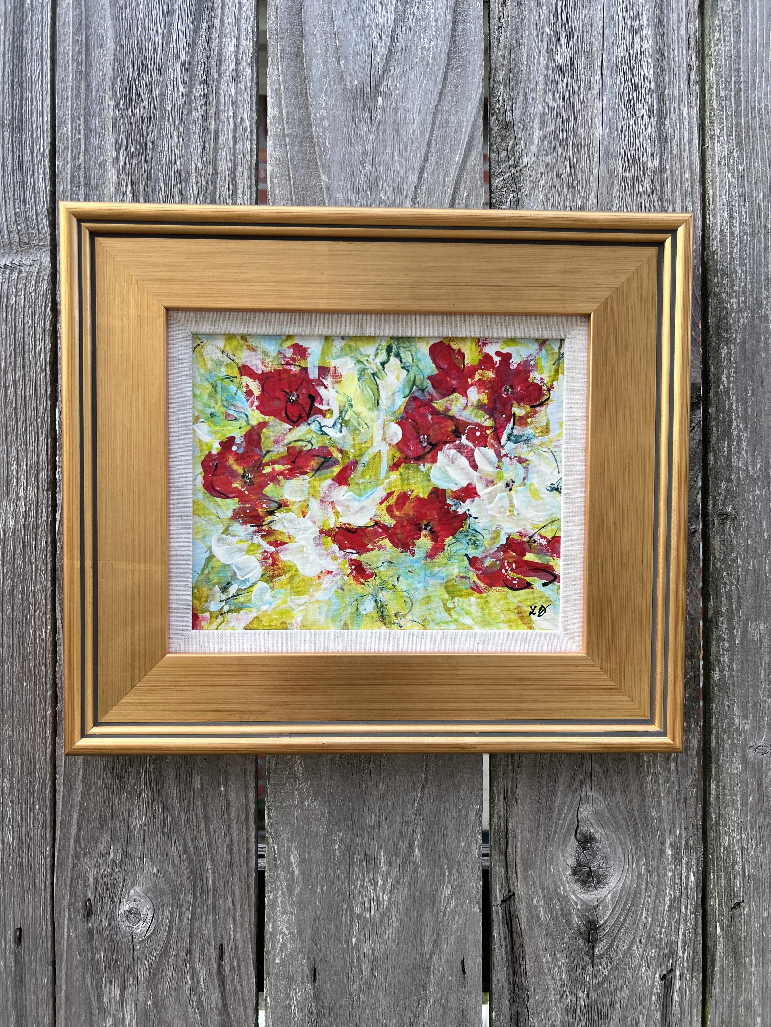 Colorful mixed media floral painting with red, white, and green expressive blooms framed in warm gold against a rustic wood backdrop.