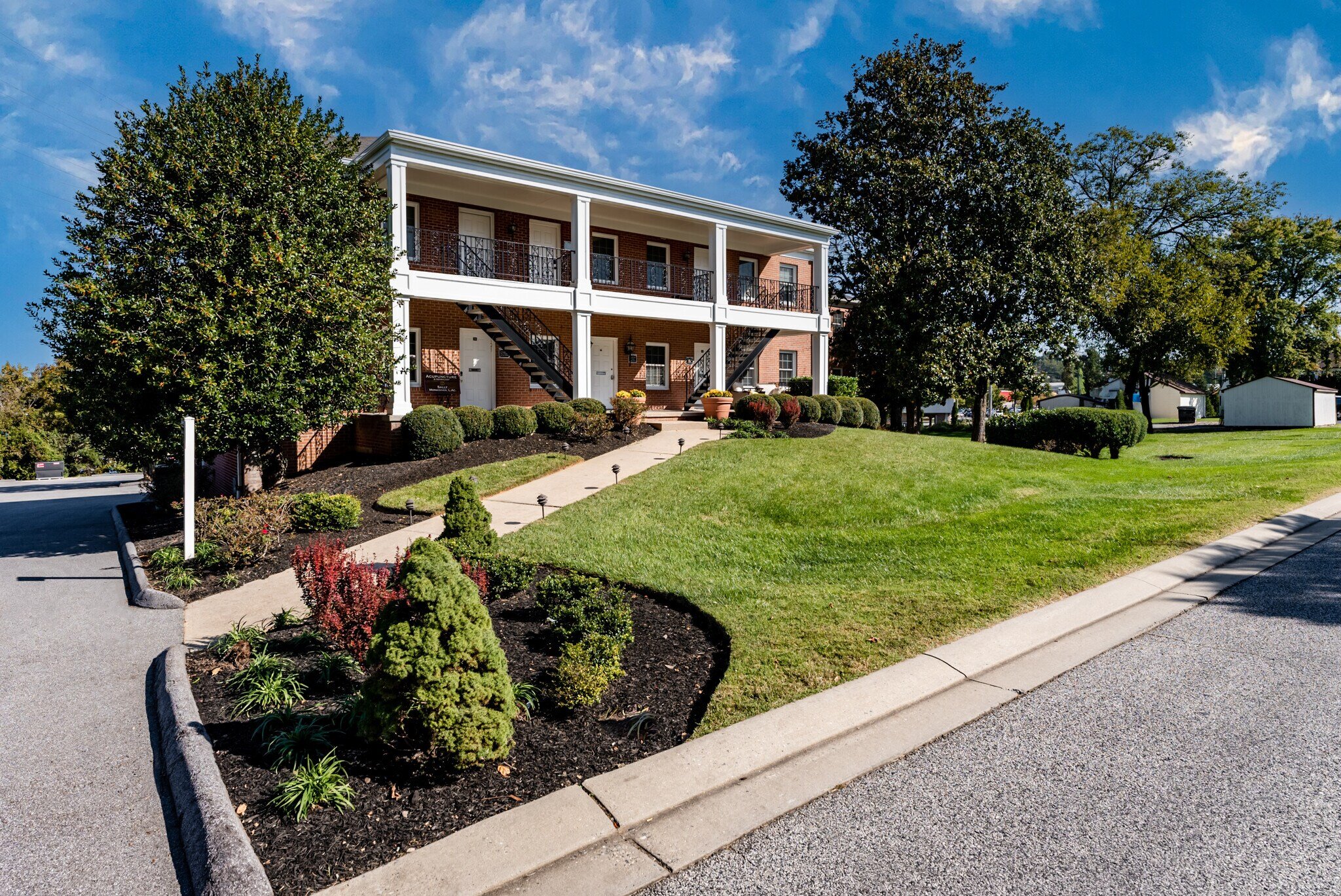 A two-story brick apartment building with white trim and black railings on the balconies, surrounded by green trees and a manicured lawn, under a partly cloudy blue sky.