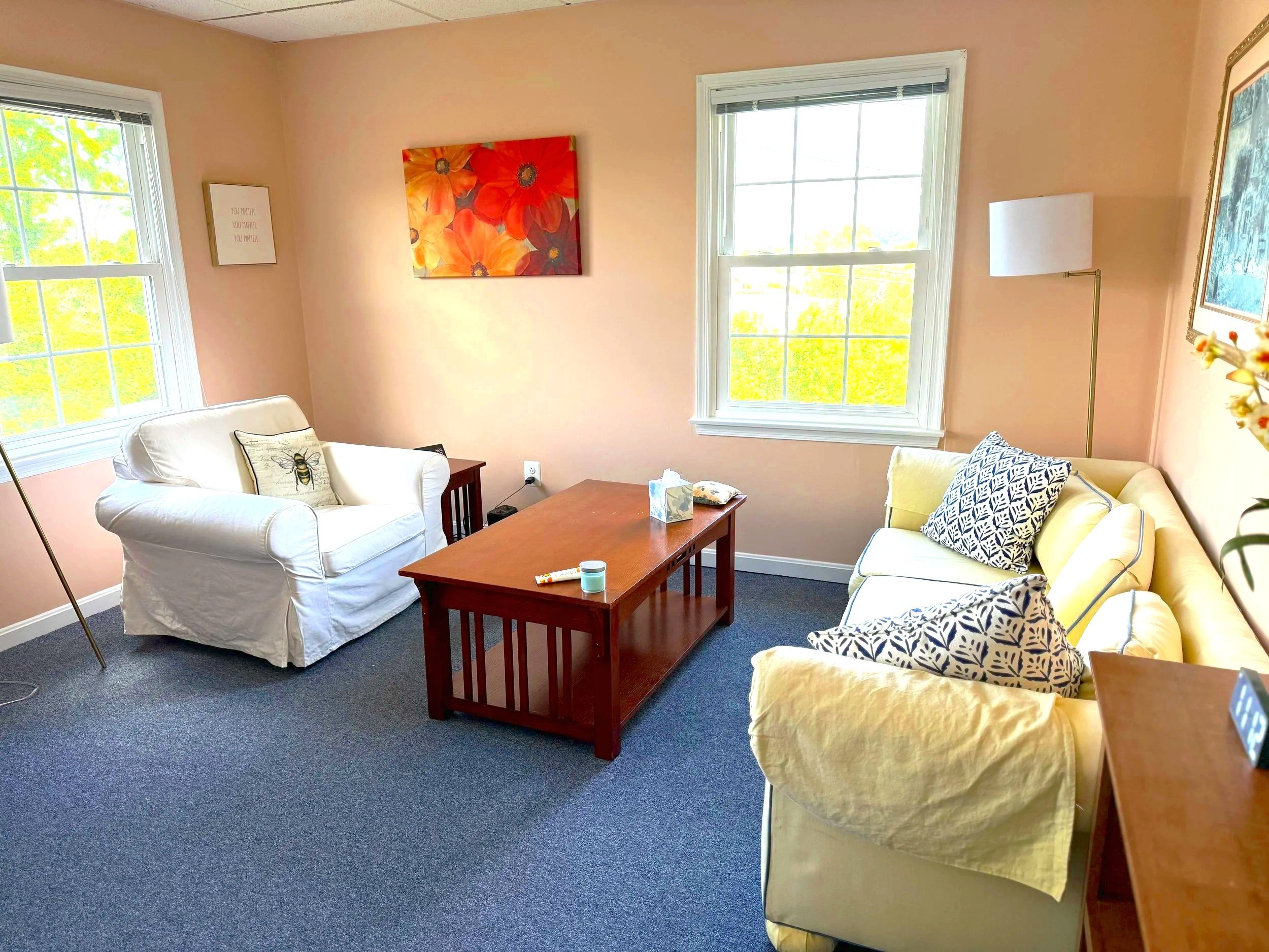 Living room with beige walls and two large windows with white trim, natural light coming in. There are two sofas with patterned and plain yellow cushions, a wooden coffee table with tissues and a small bottle on it, and a standing lamp. There are paintings on the wall, one floral and one abstract, and a small framed quote. The carpet is dark-colored.