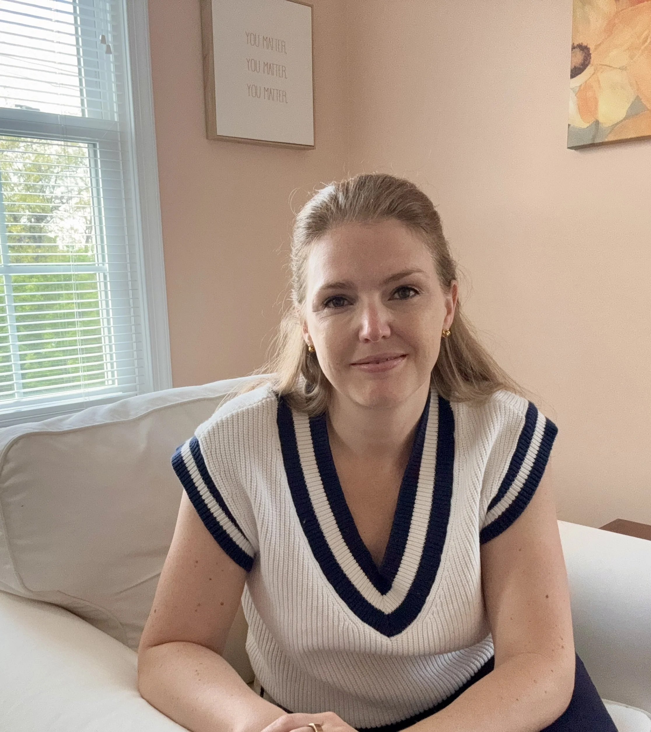 A woman with blonde hair, wearing gold earrings and a white knit top with navy stripes, is sitting on a beige couch in a room with light pink walls. Behind her, there is a framed quote and a floral painting on the wall.  Erin Birely, LCPC