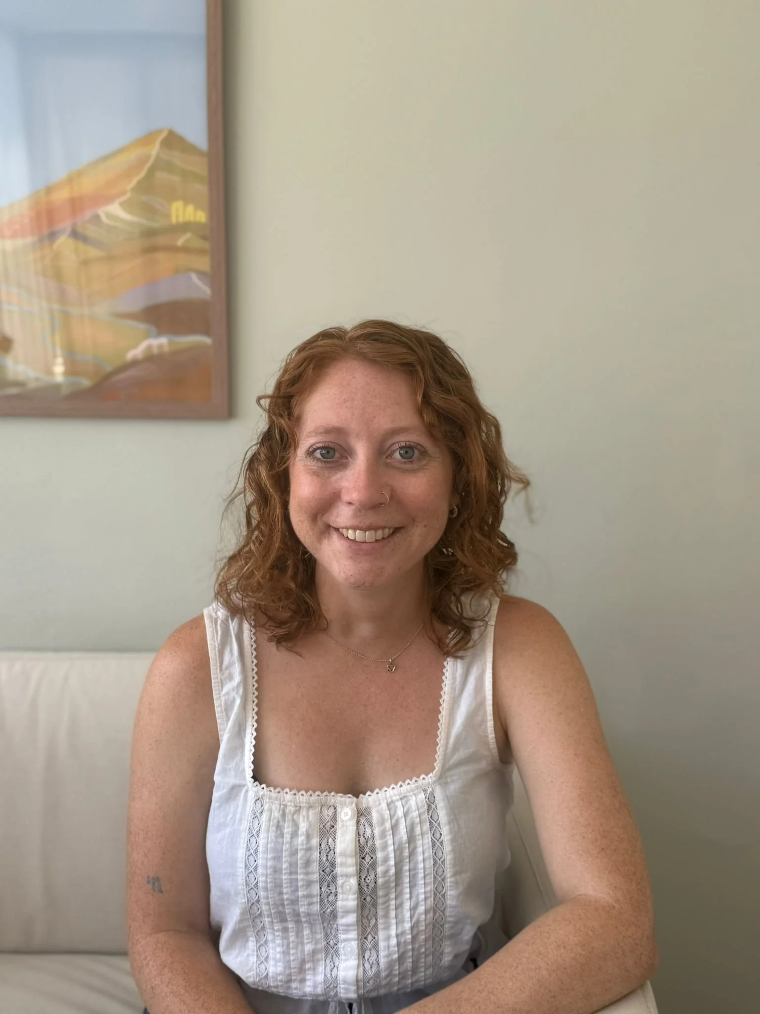 A woman with curly red hair smiling, wearing a white sleeveless top, sitting in front of a light-colored wall with landscape artwork visible in the background. Elizabeth Foote, LCPC