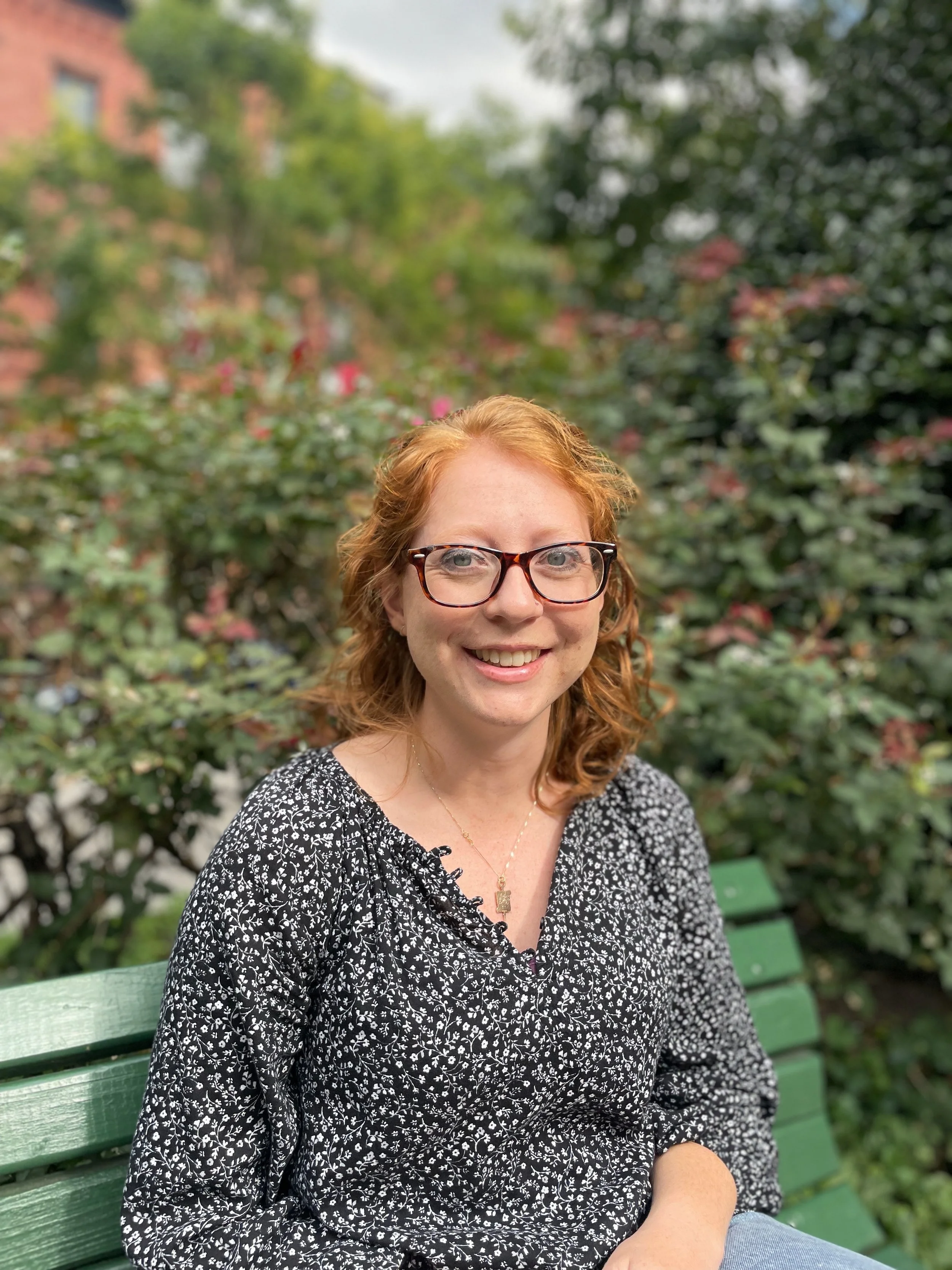 A smiling woman with red hair, wearing glasses and a black and white floral top, sitting on a green park bench with lush green trees and pink flowers in the background. Elizabeth Foote, LCPC