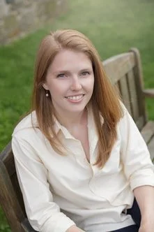 A young woman with red hair, smiling, sitting on a wooden bench outdoors with green grass in the background. Erin Birely, LCPC