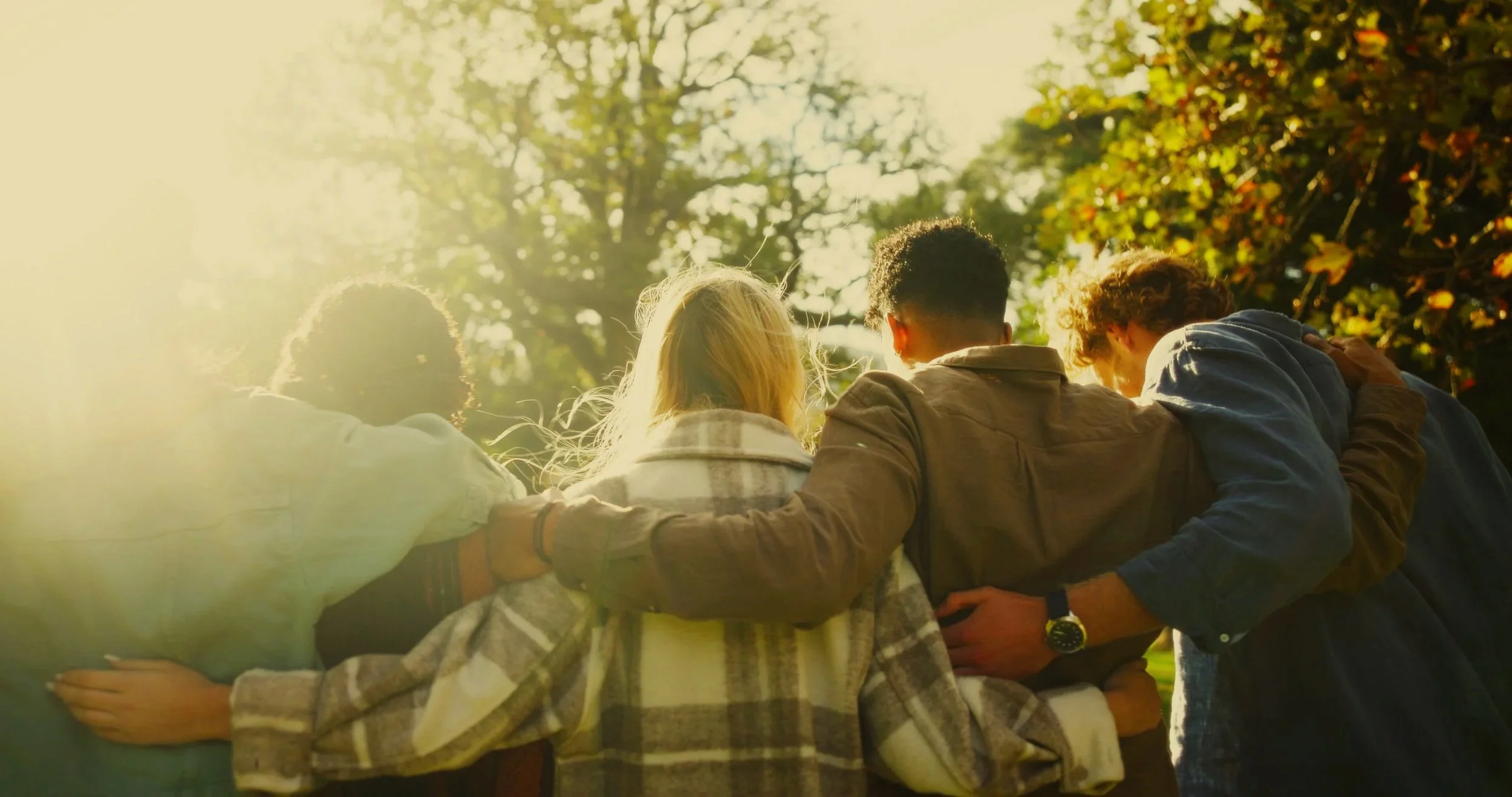 A group of young people sitting outdoors in a circle with their arms around each other, facing away from the camera on a sunny day in a park with trees.