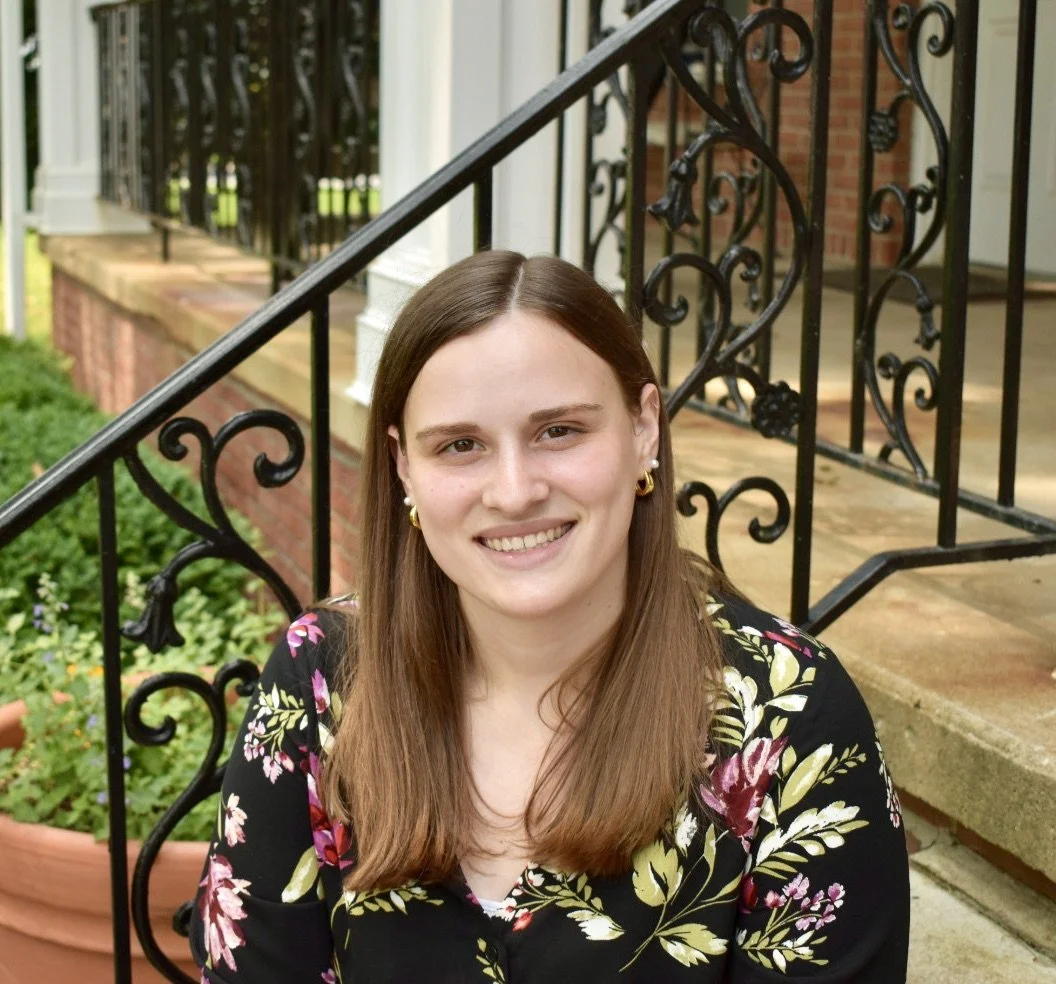 A woman with long brown hair, wearing a black floral top and gold hoop earrings, smiling outdoors near a brick house with black wrought iron railings, green plants, and potted flowers. Savannah Blythe, LCPC
