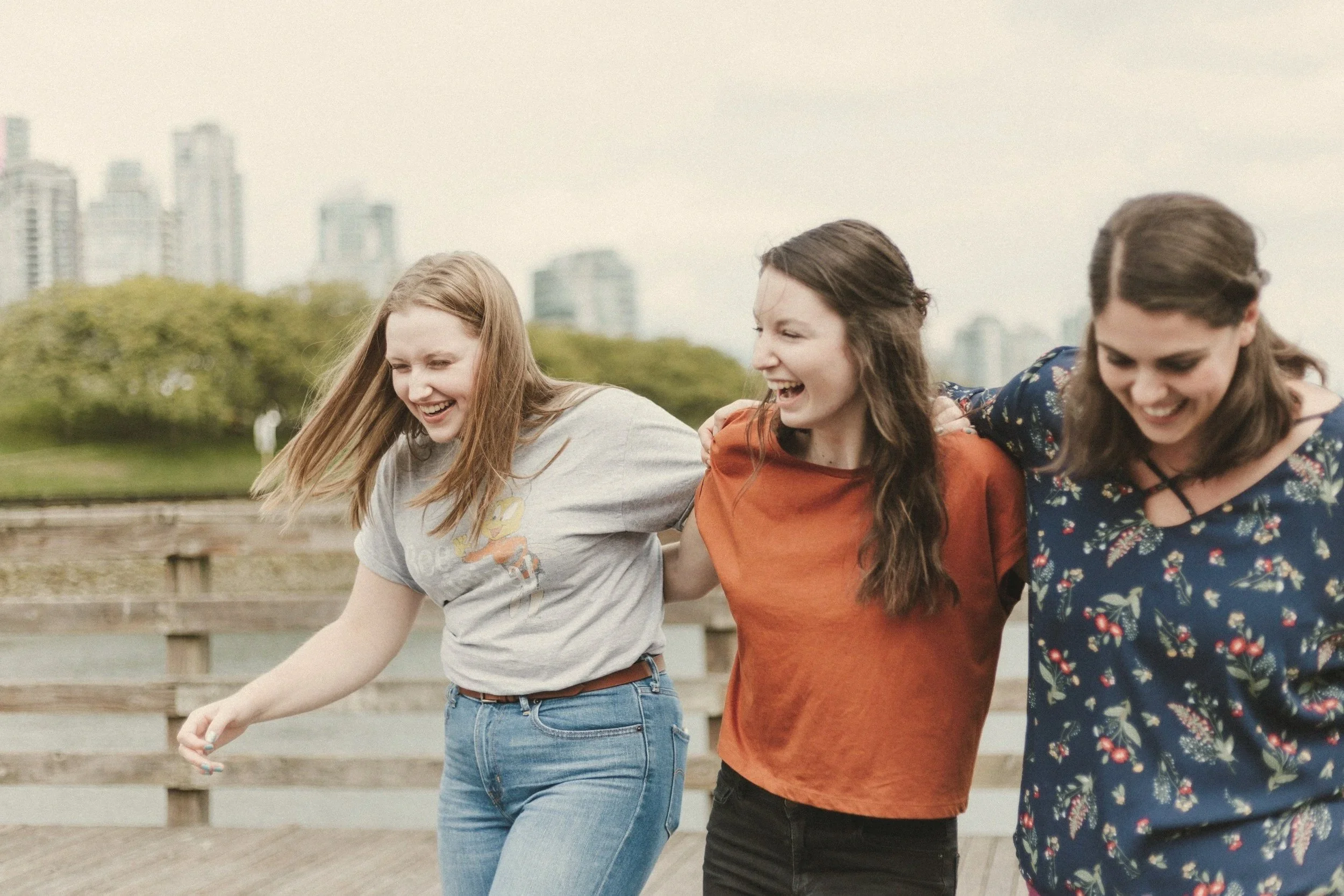 Three young women smiling and holding each other as they walk outdoors near a park with city buildings in the background.