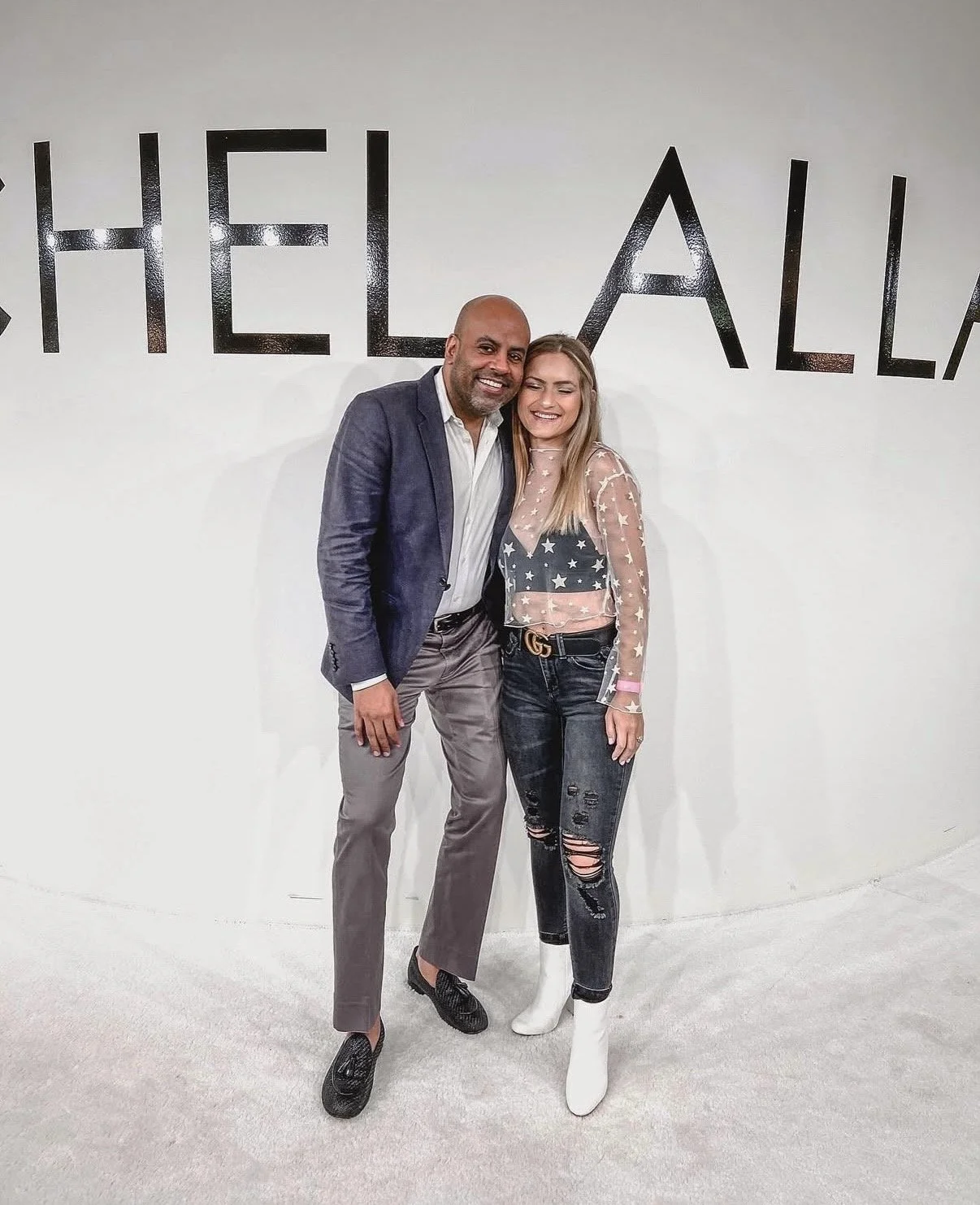 A man and woman standing together, smiling, in front of a wall with partially visible black text. The woman is wearing white boots, distressed jeans, and a star-patterned sheer top. The man is in a blazer and dress shoes.