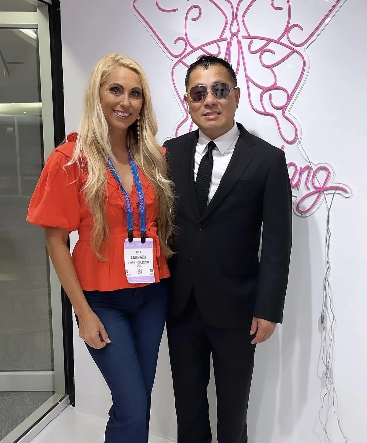 A woman in a bright orange ruffled top and dark blue jeans standing next to a man in a black suit, white shirt, and black tie, both smiling. The woman has long blonde hair and is wearing earrings, with a conference badge hanging around her neck. Behi
