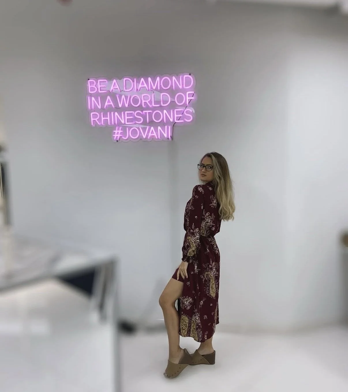 A woman with glasses and long blonde hair wearing a burgundy dress with a floral pattern, standing against a white wall. A pink neon sign reads, 'BE A DIAMOND IN A WORLD OF RHINESTONES #JOVANI'.