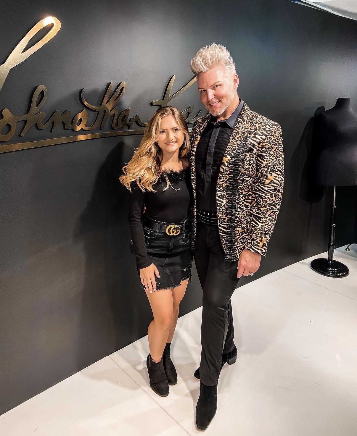 A man and a woman standing indoors in front of a black wall with a gold signature logo. The woman has long blond hair, wearing a black long-sleeve top, a black denim skirt, and black ankle boots. The man has platinum blond hair, wearing a leopard pri