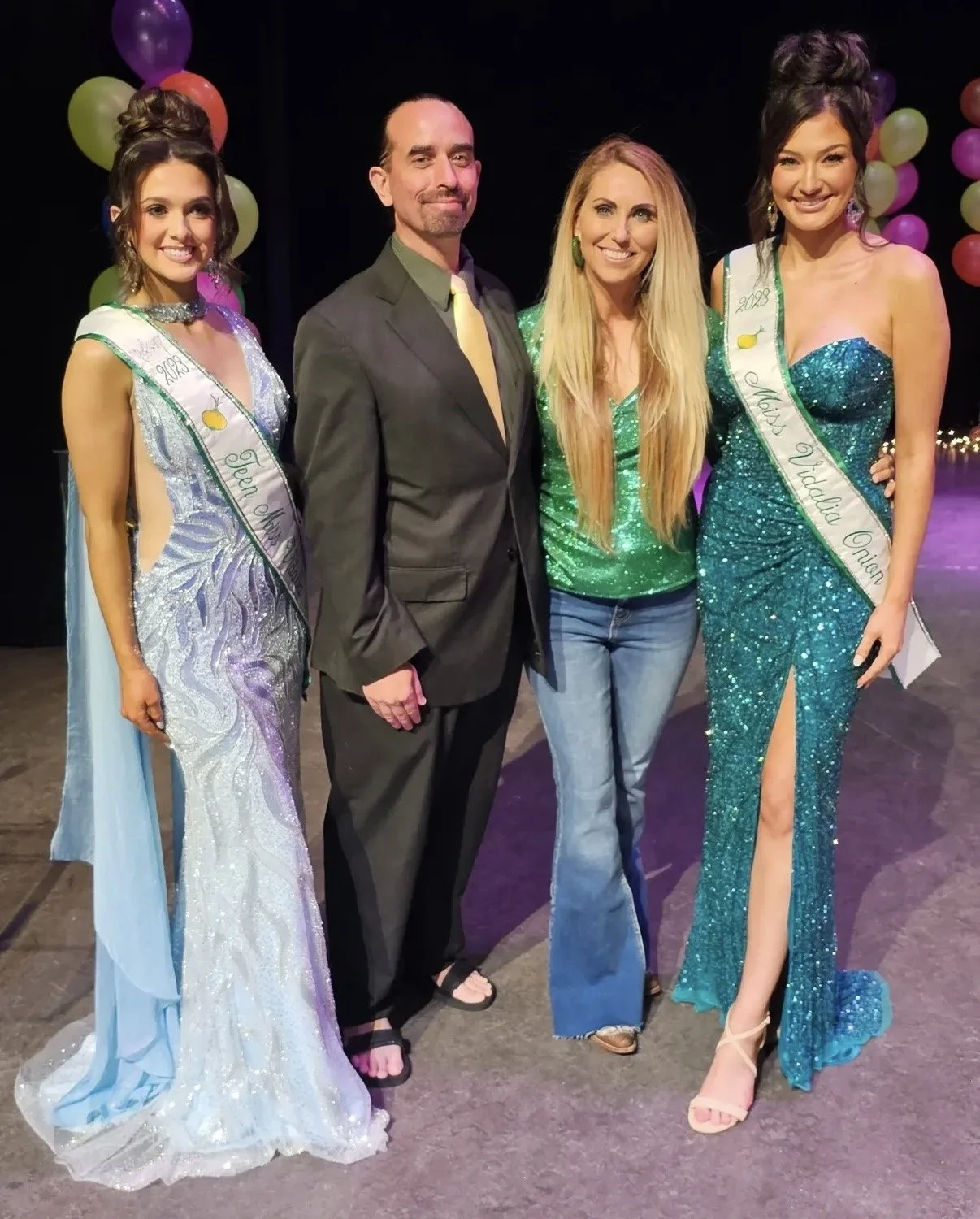 Two women dressed in evening gowns and two men in suits, standing on a stage decorated with colorful balloons, during a pageant or celebration.