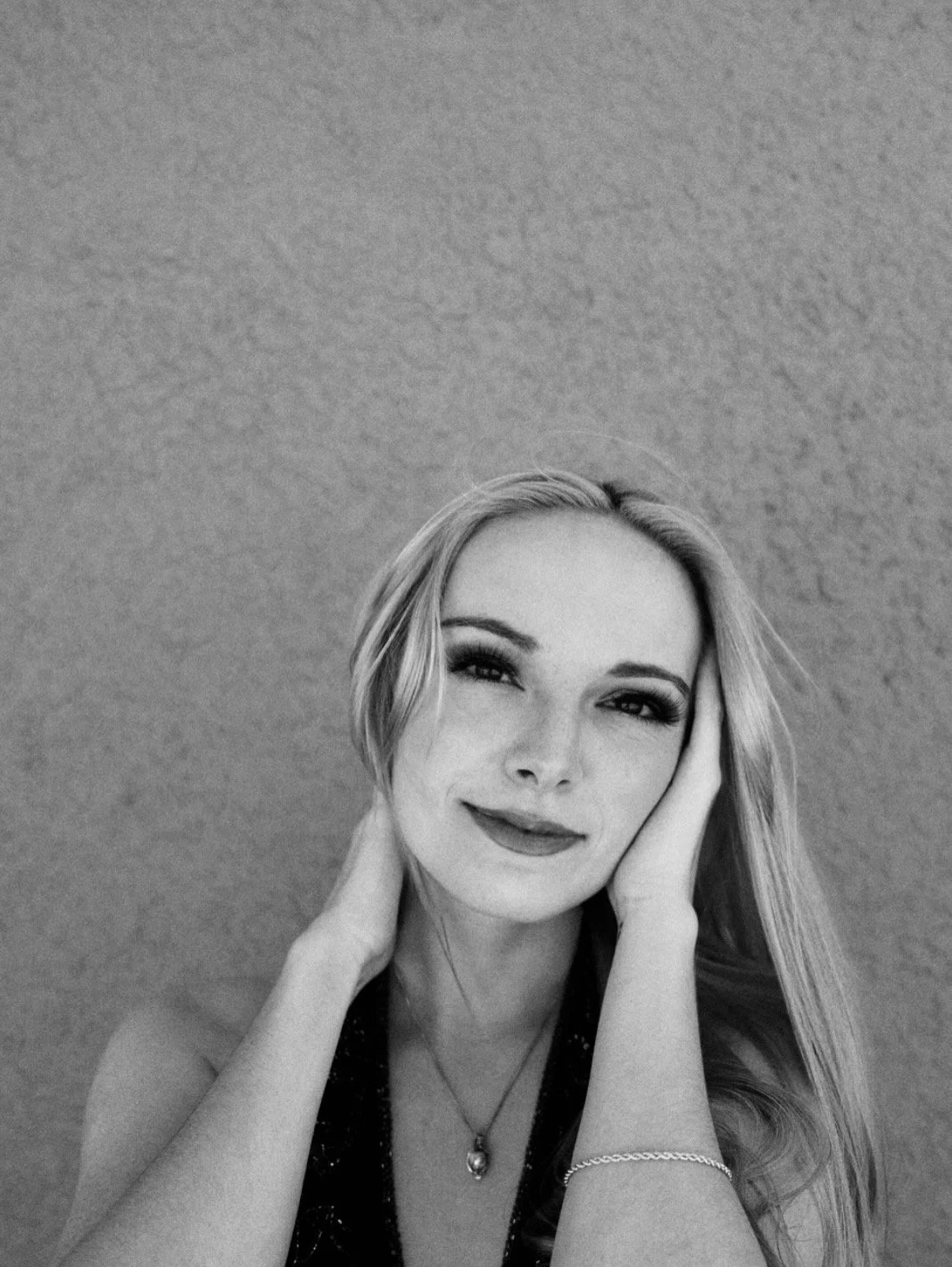 Black and white portrait of a woman with long hair, smiling gently, with her hands touching the sides of her face, against a plain background.