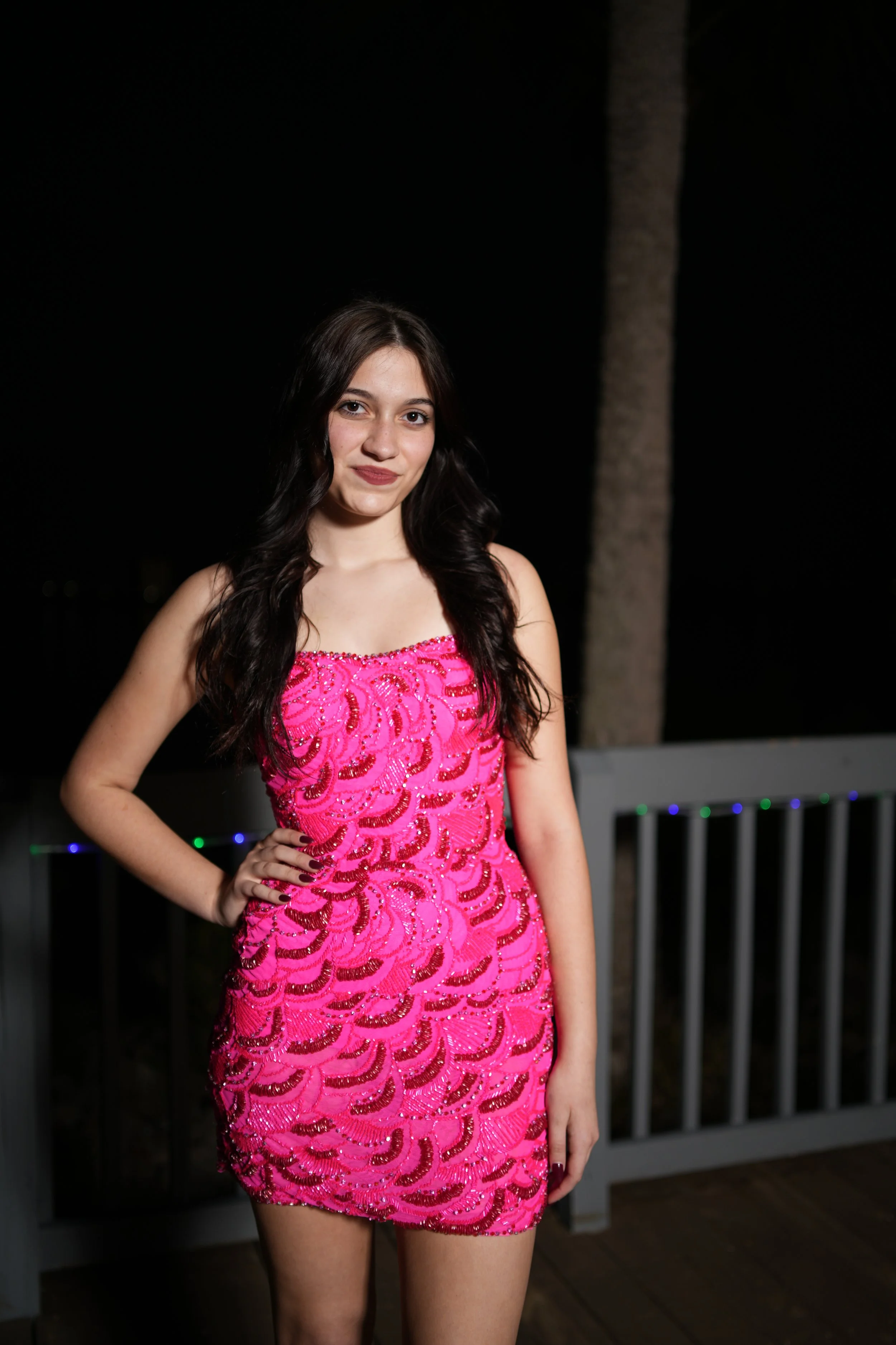 A young woman with long dark hair wearing a pink sequined dress with a swirling pattern, standing outdoors at night next to a wooden railing and a tree.