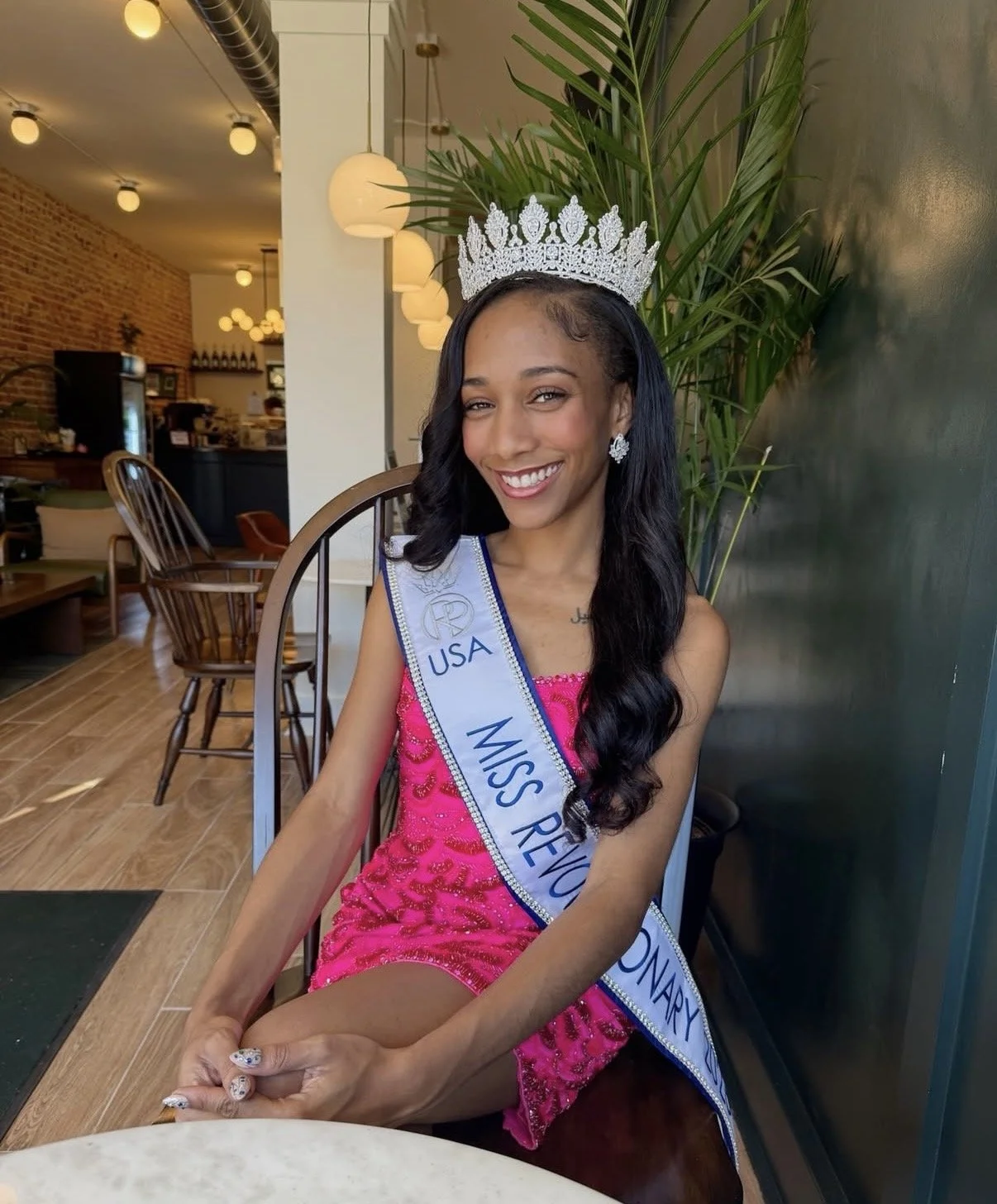 Young woman wearing a crown and sash reading Miss Revi County, smiling, sitting in a cafe setting.