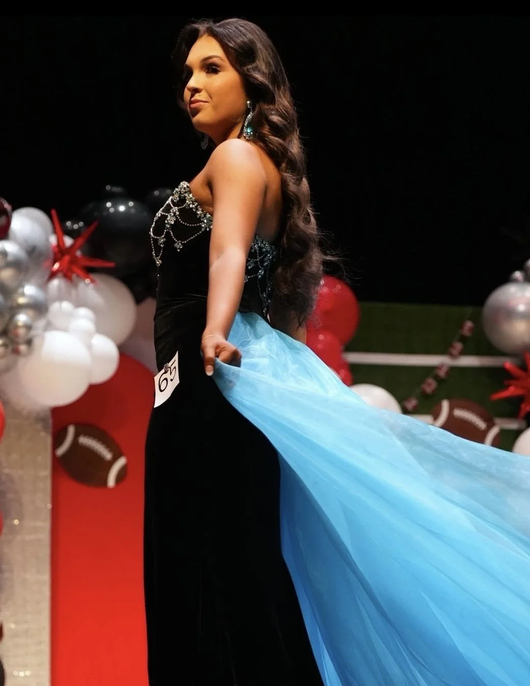 A woman on stage participating in a pageant or fashion show, wearing a black dress with embellishments and a large blue tulle train, with decorative balloons and festive decorations in the background.