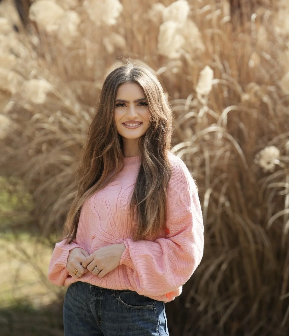 A young woman with long wavy brown hair, wearing a pink sweatshirt and jeans, standing outdoors in front of tall, dried grass or wheat, smiling at the camera.