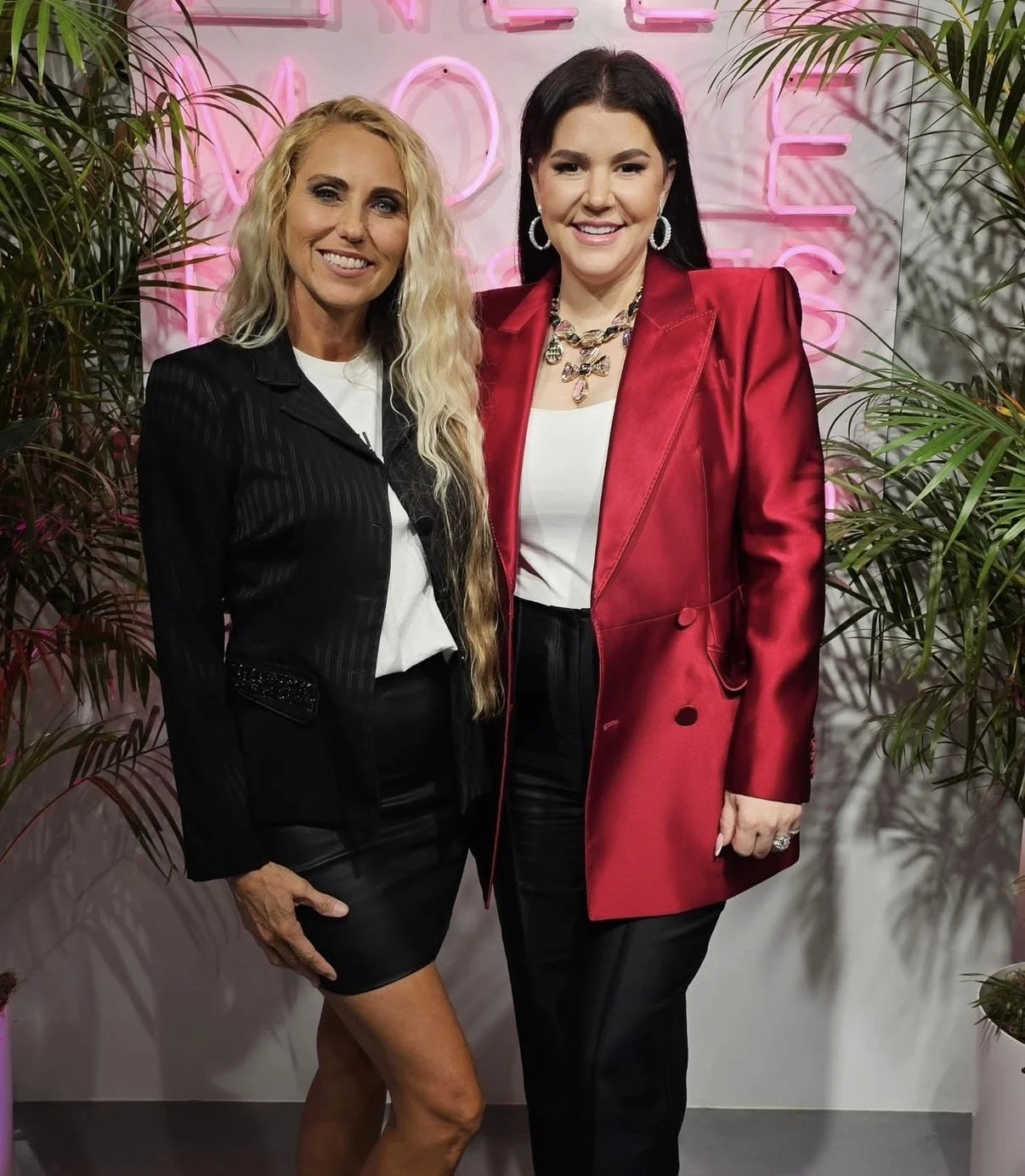 Two women standing side by side in front of a pink neon sign that reads 'LOVE' with greenery on either side, both smiling and dressed in stylish clothing.