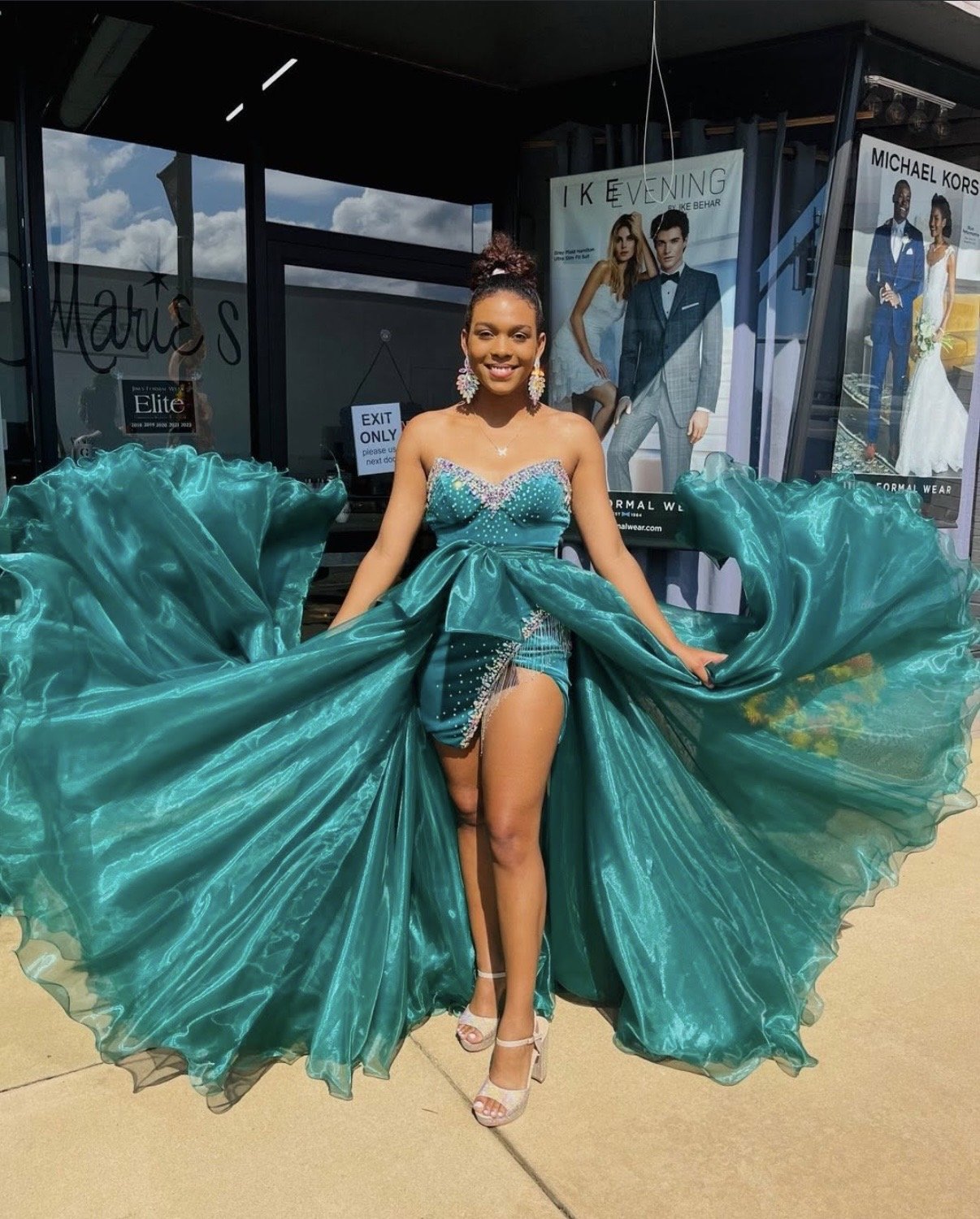 A woman in a teal dress with a voluminous skirt poses outside a store, lifting the skirt to reveal her legs. The dress is shiny with embellishments at the top. She is smiling, wearing earrings, a necklace, and heels. Behind her are posters of formal 
