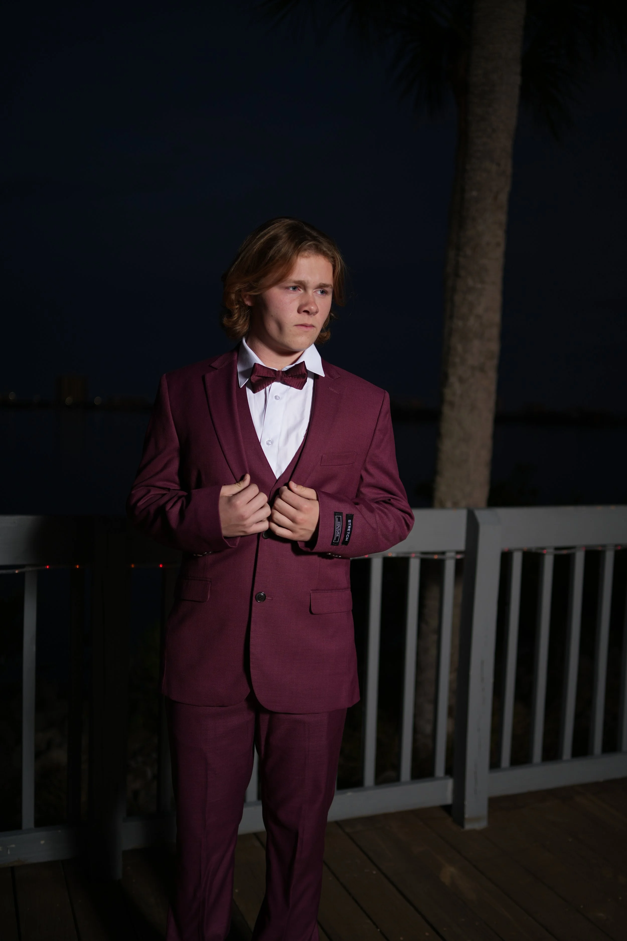 A young man with long blonde hair in a burgundy suit and bow tie standing on a balcony at night near a palm tree, with a body of water in the background.