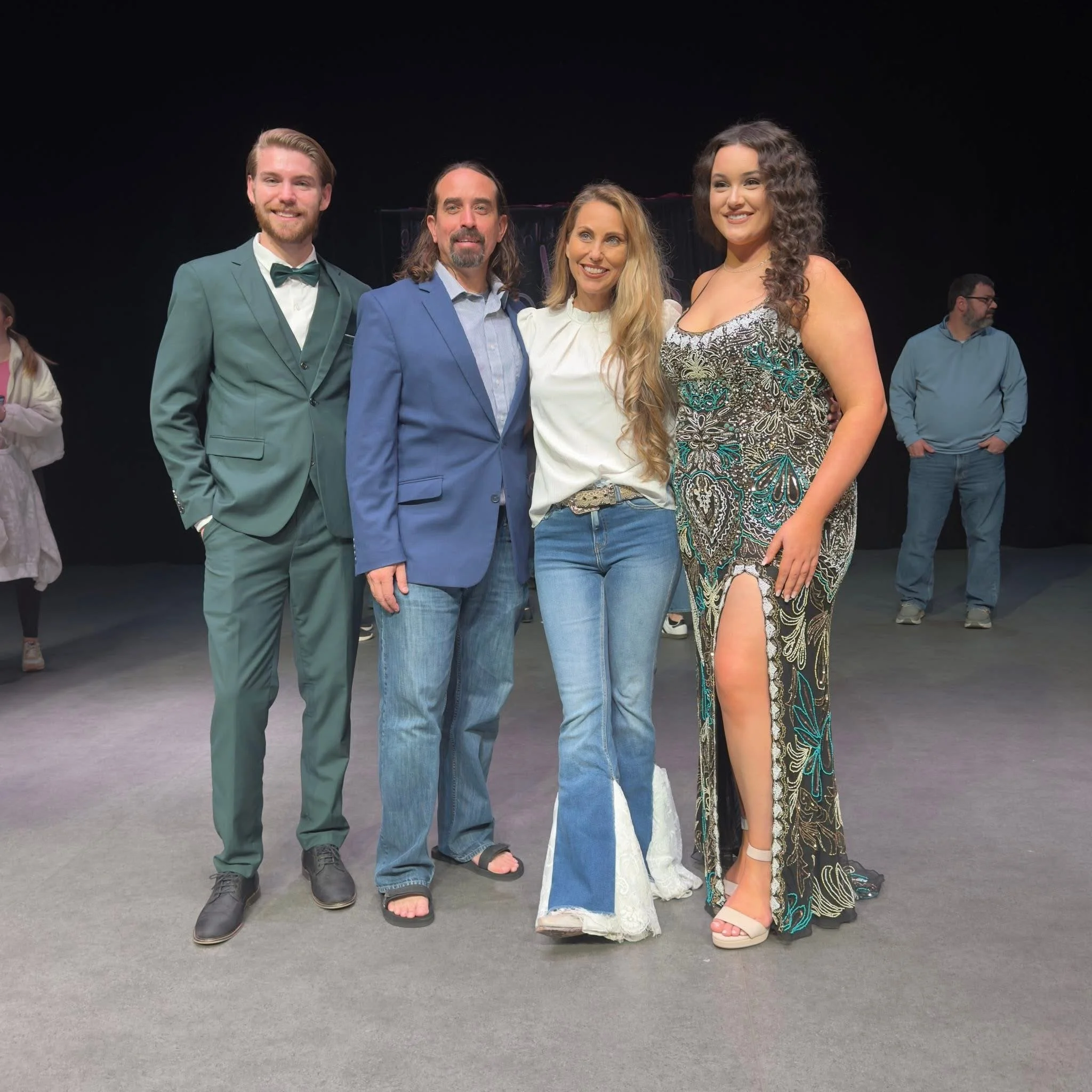 Group of five people standing together on a stage, four facing forward and smiling, one in the background looking to the side. Two men and three women dressed in formal and casual attire.