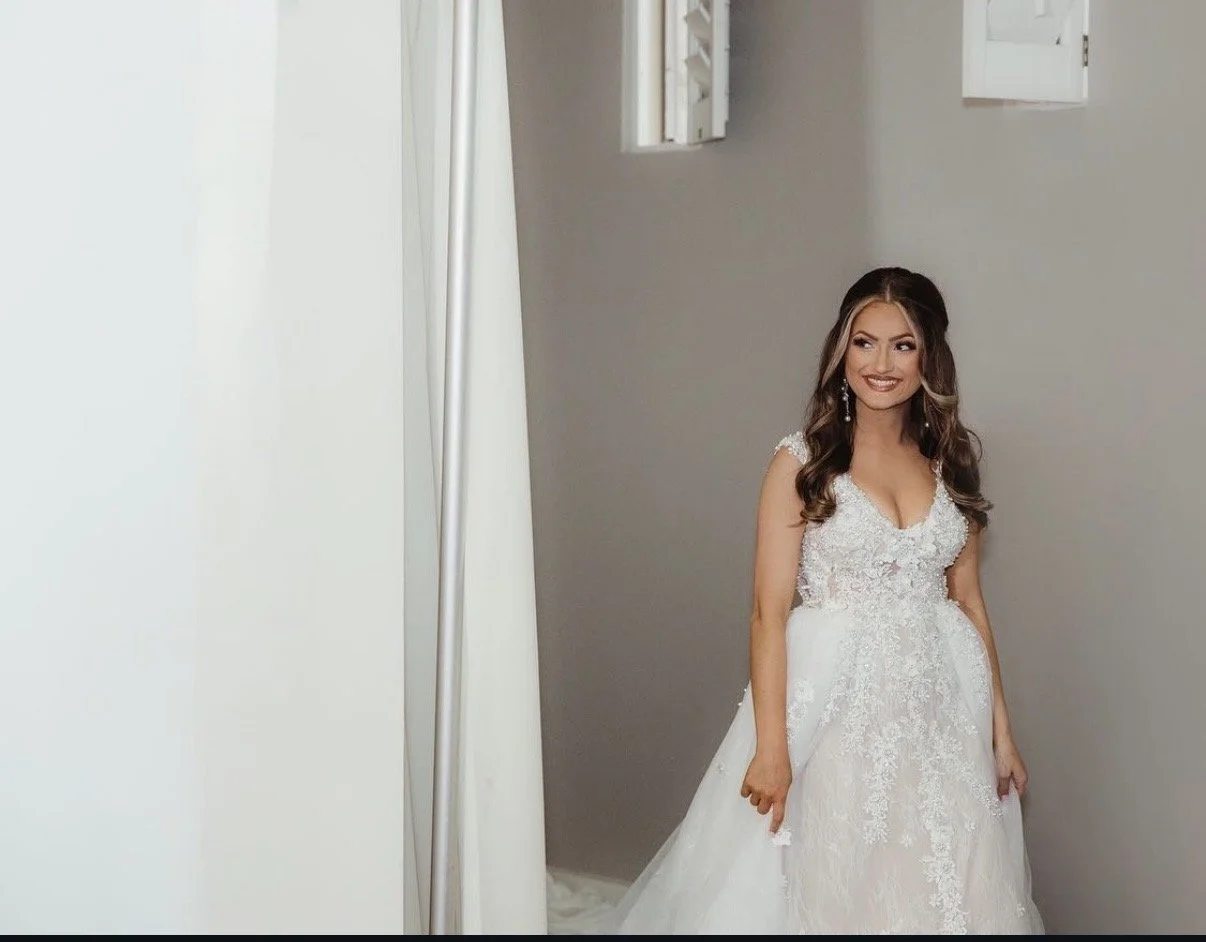 A woman in a wedding dress standing indoors in front of a gray wall, smiling.